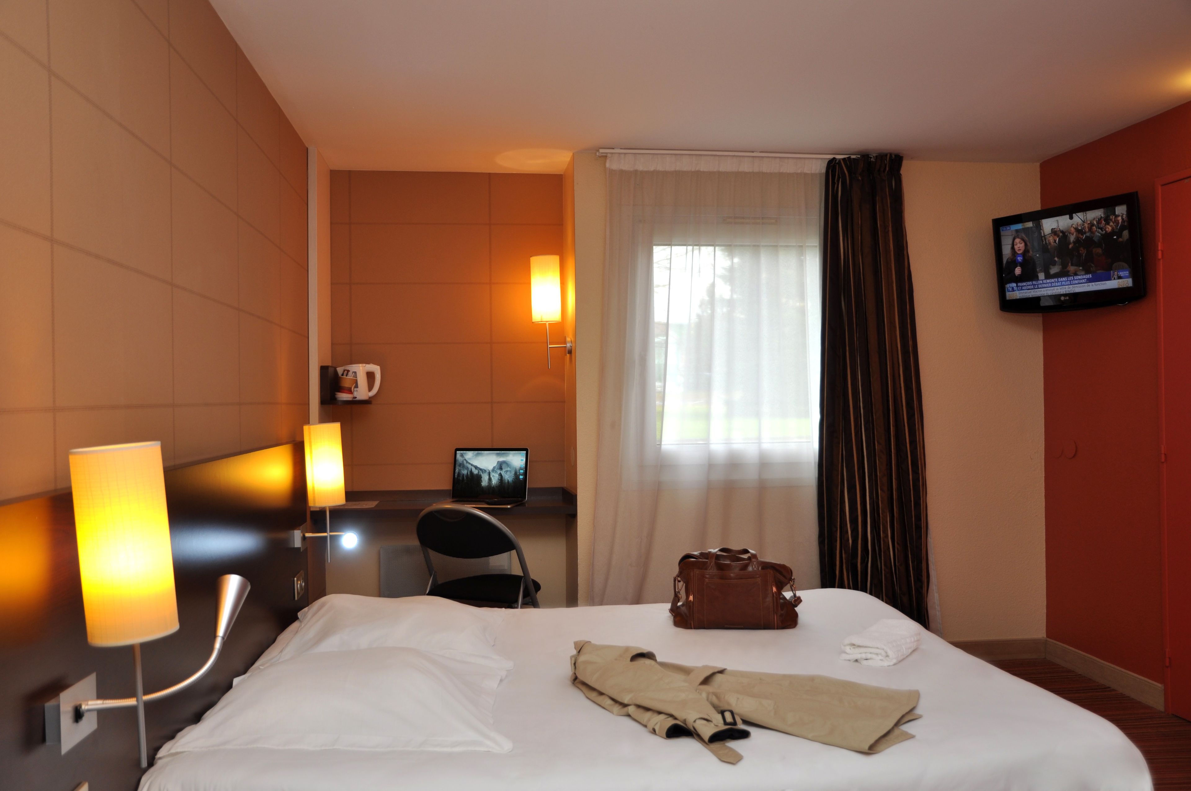 Een hotelkamer met een bed, een bureau met een computer, een televisie aan de muur en een raam met gordijnen.