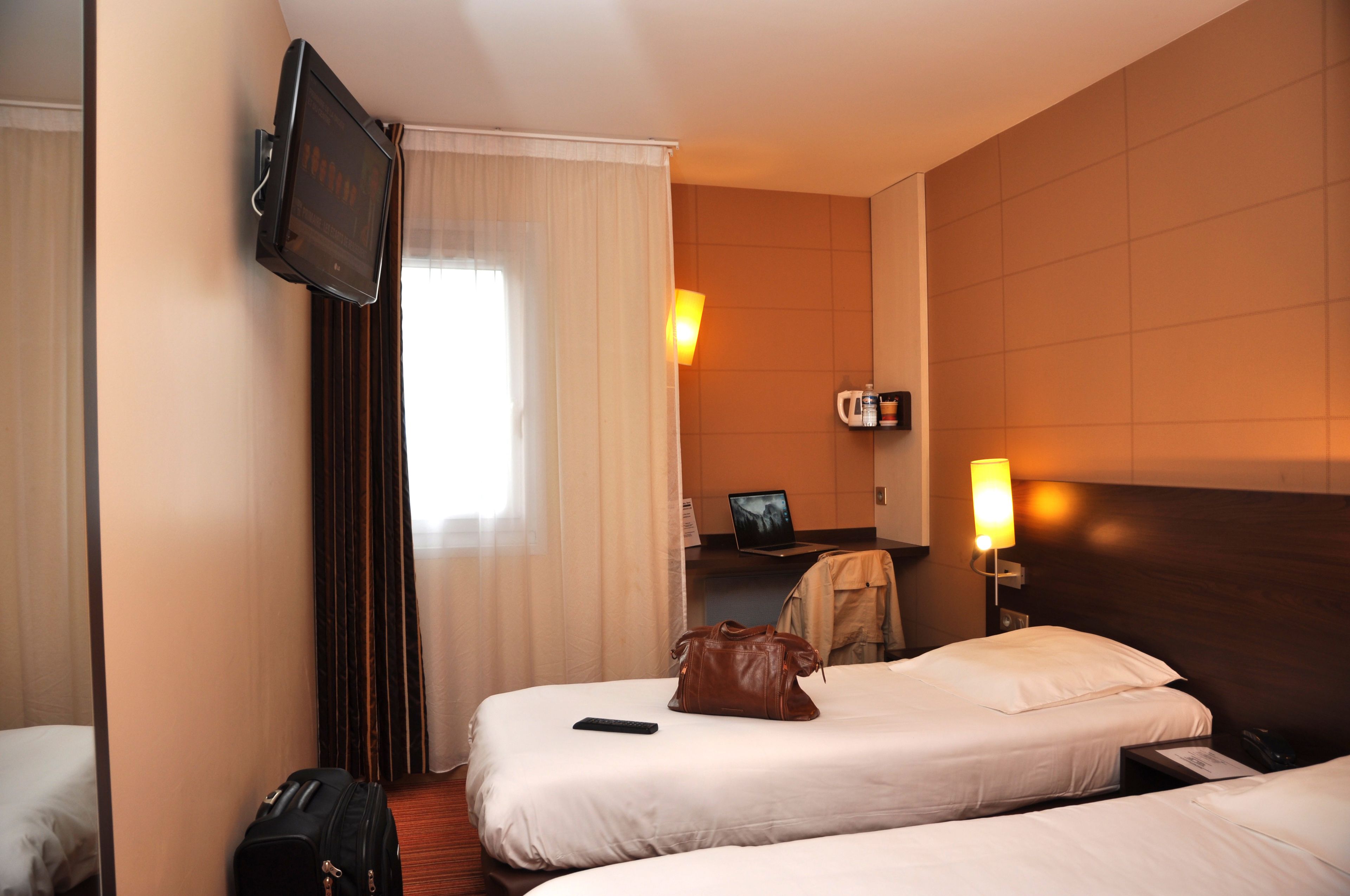 Een hotelkamer met twee bedden, een aan de muur bevestigde televisie, een bureau met een laptop en een lamp.