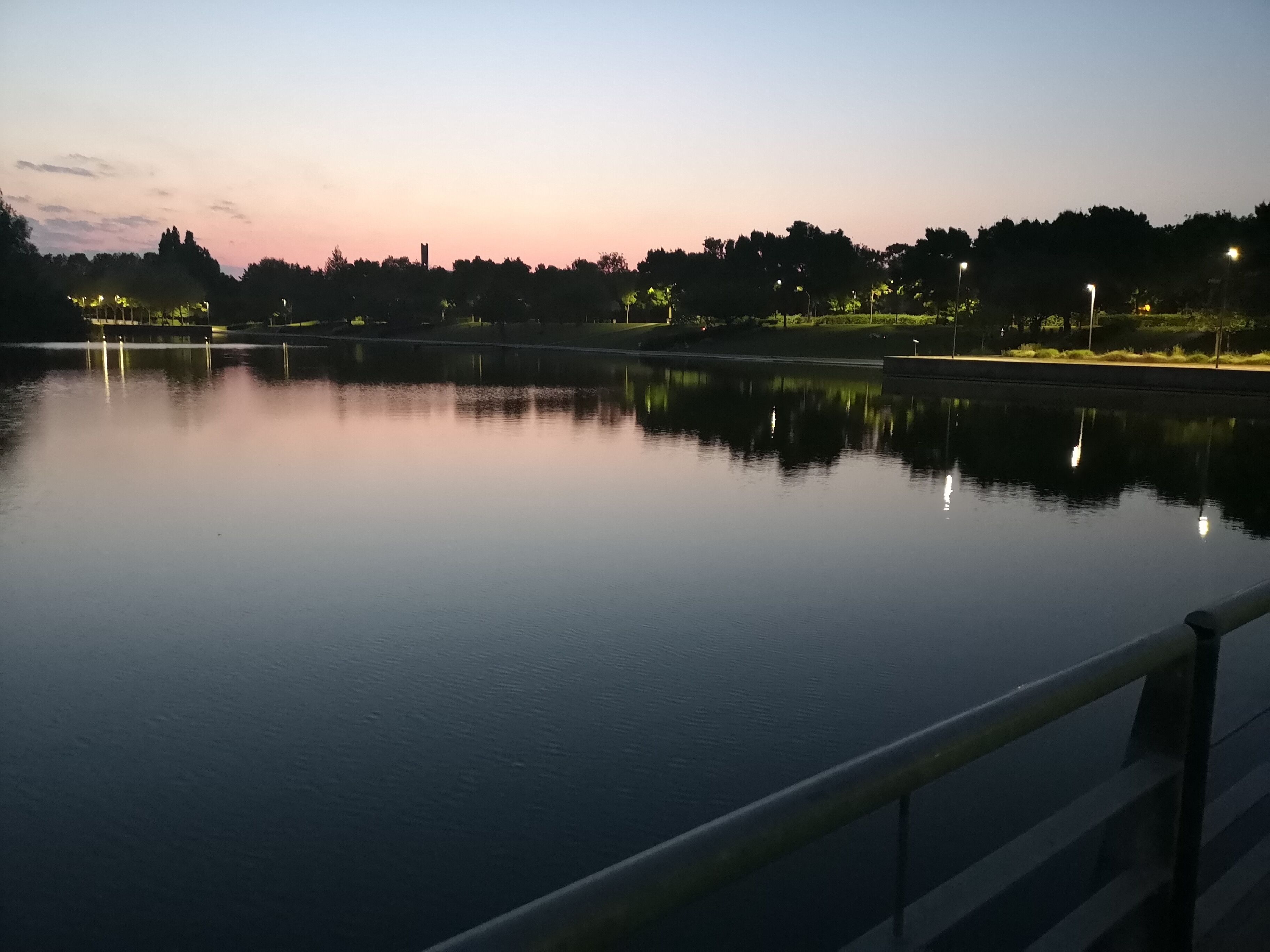 A calm lake at dusk with a clear sky, a distant shoreline with trees, and a railing in the foreground.