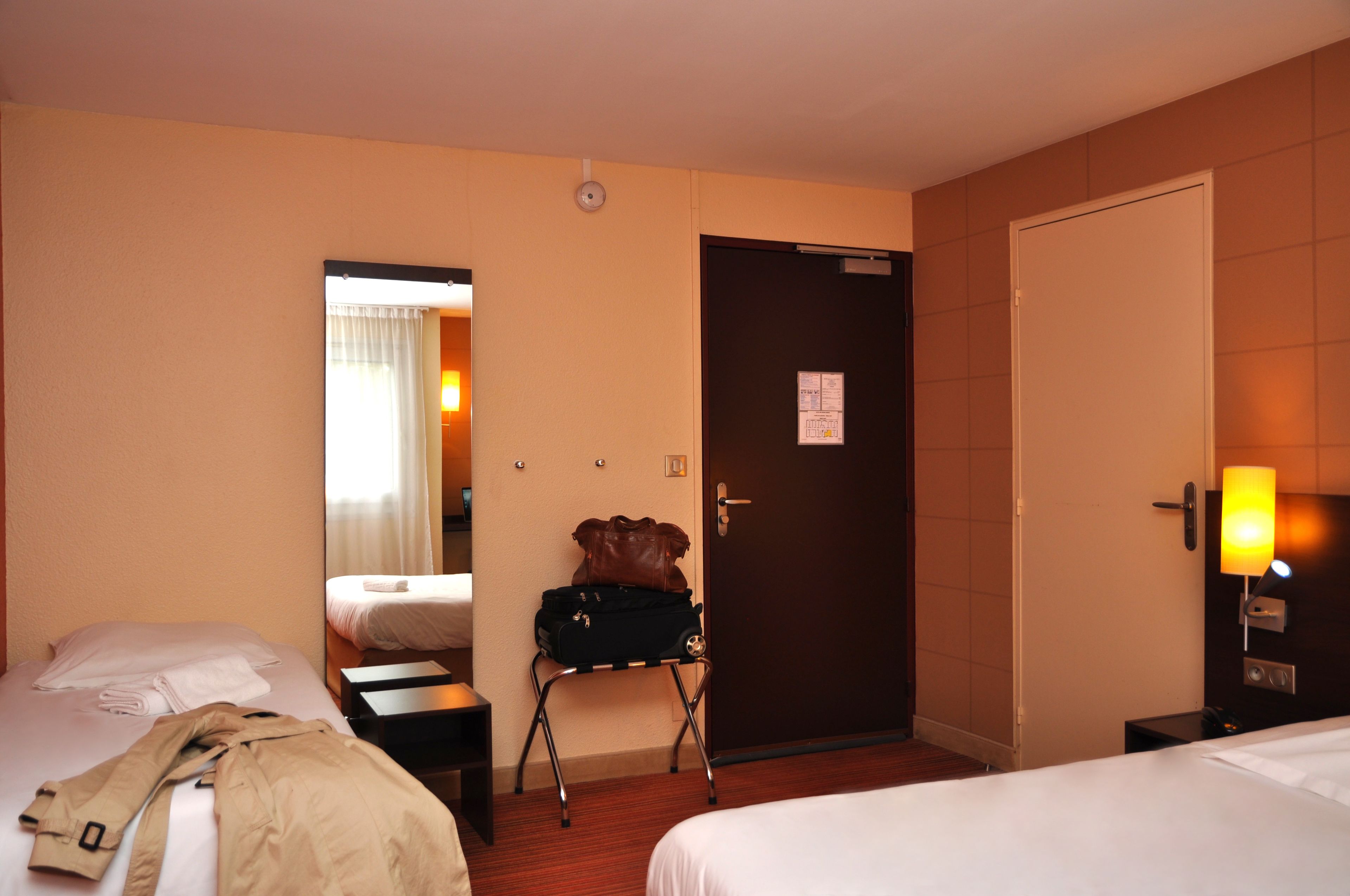 Habitación de hotel con dos camas, una maleta sobre una silla, una puerta y una mesita de noche con una lámpara.