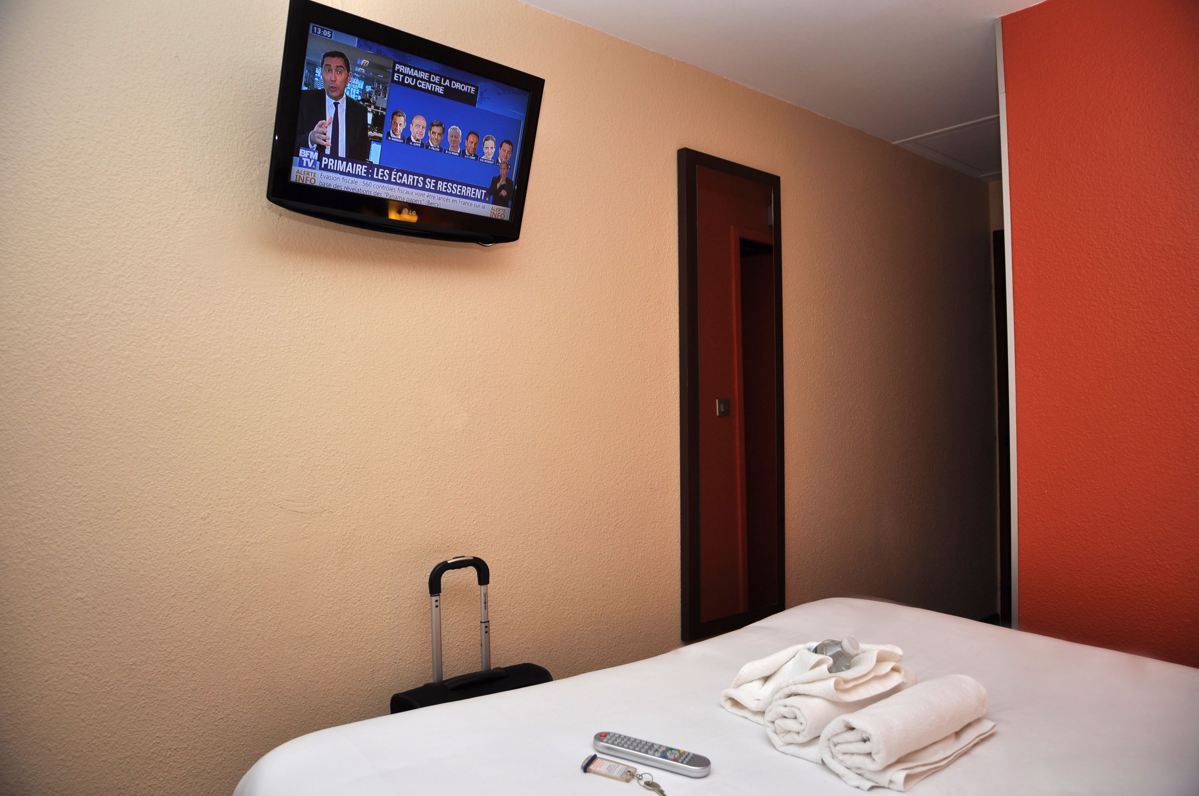 Ein Hotelzimmer mit einem Bett, einem an der Wand befestigten Fernseher, der eine Nachrichtensendung zeigt, einem Koffer und einer Fernbedienung auf dem Bett.