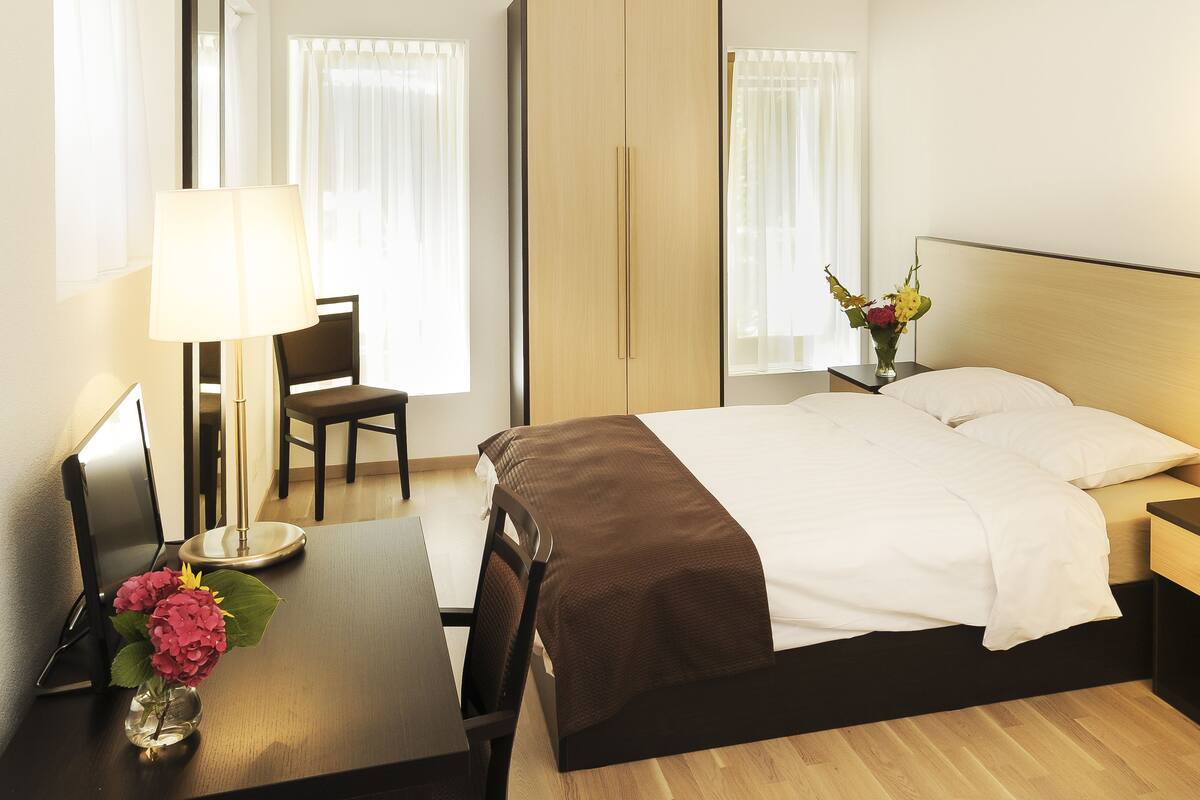 Ein Hotelzimmer mit einem Bett, Nachttischen, einem Schreibtisch mit Lampe, einem Stuhl und einem Blumenstrauß.