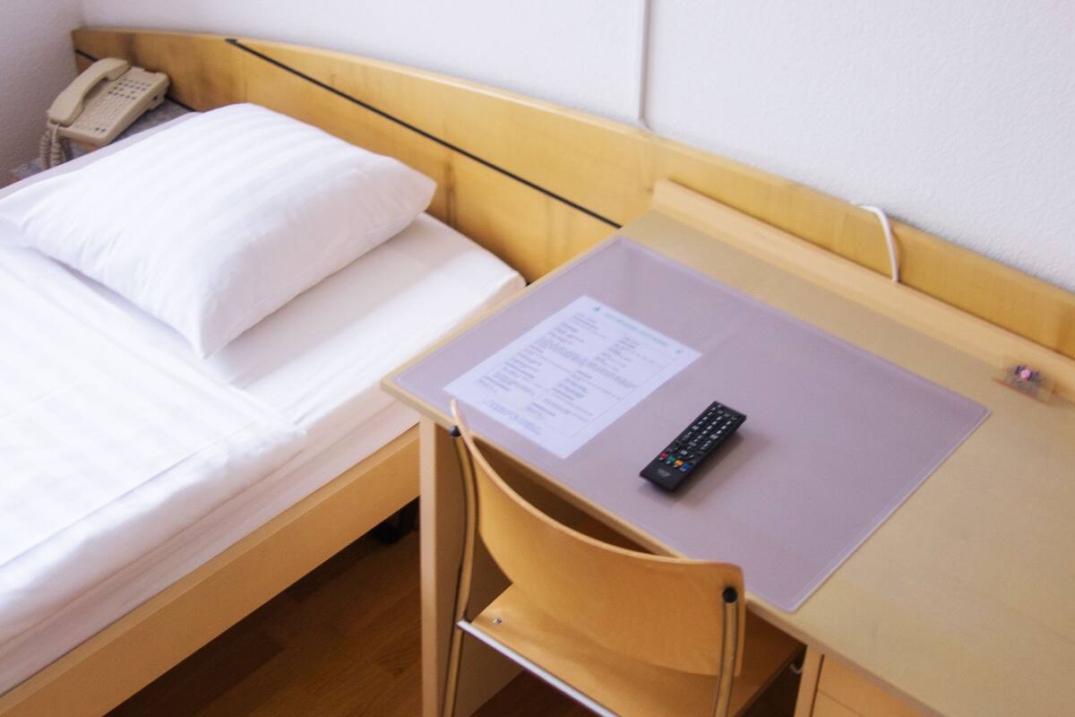 Ein Einzelbett mit weißer Bettwäsche, ein Nachttischtelefon, ein Schreibtisch mit Fernbedienung und ein Sessel.