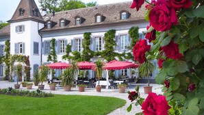 Un bâtiment à la façade blanche, doté de nombreuses fenêtres et surmonté d’une tour. On y trouve des tables à l’extérieur, protégées par des parasols rouges, le tout étant entouré de plantes en pot et d’une pelouse bien entretenue.