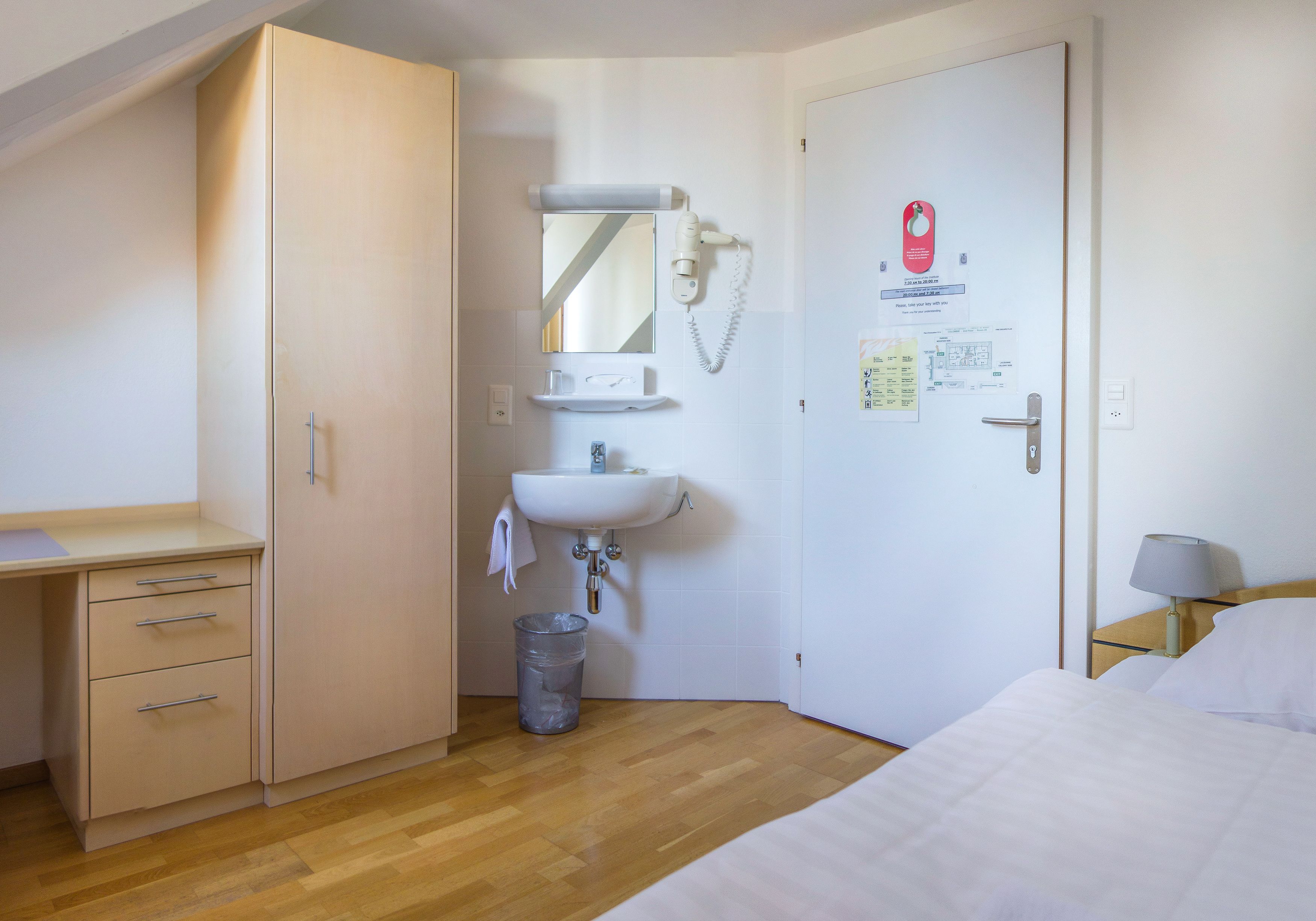 A hotel room with a wooden floor, a bed with white bedding, a bedside table with a lamp, a wardrobe, a sink with a mirror, and a trash bin.