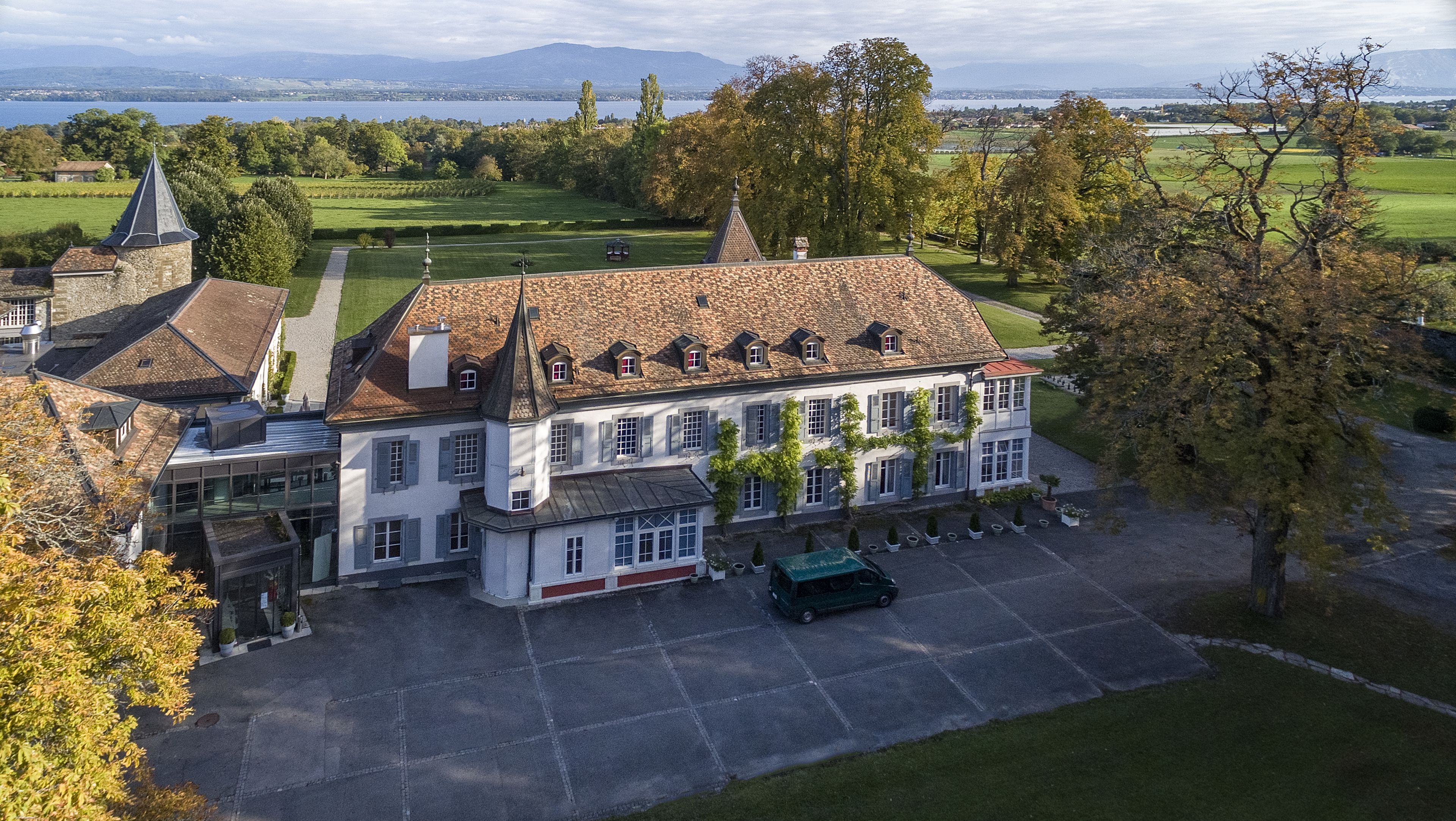 Un grand bâtiment à plusieurs étages, doté d’une tour imposante, d’une cour intérieure et offrant une vue sur un lac et des montagnes en arrière-plan.