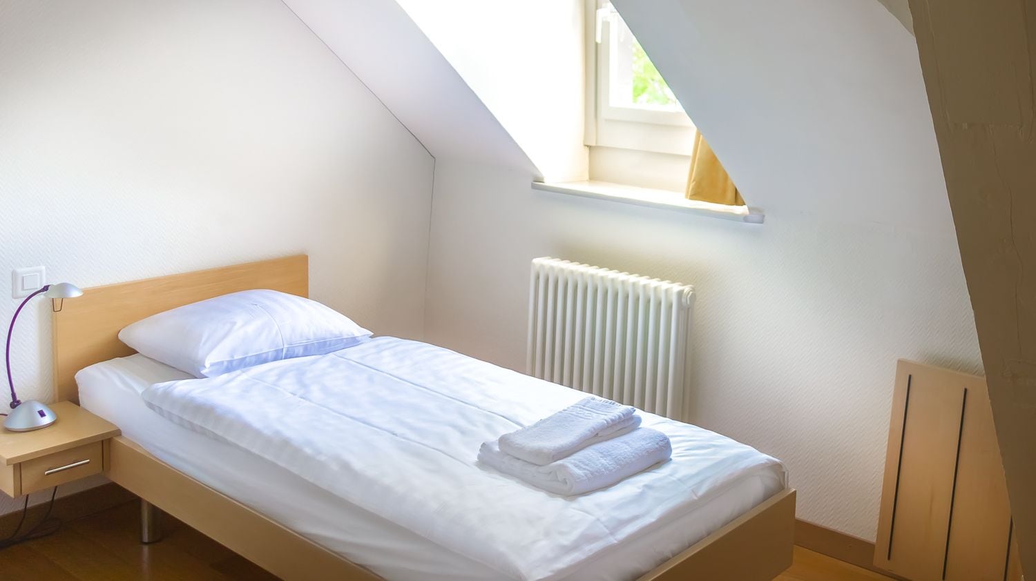 Una cama individual con ropa de cama blanca, una mesita de noche con lámpara, una ventana con vistas a zonas verdes y un radiador.