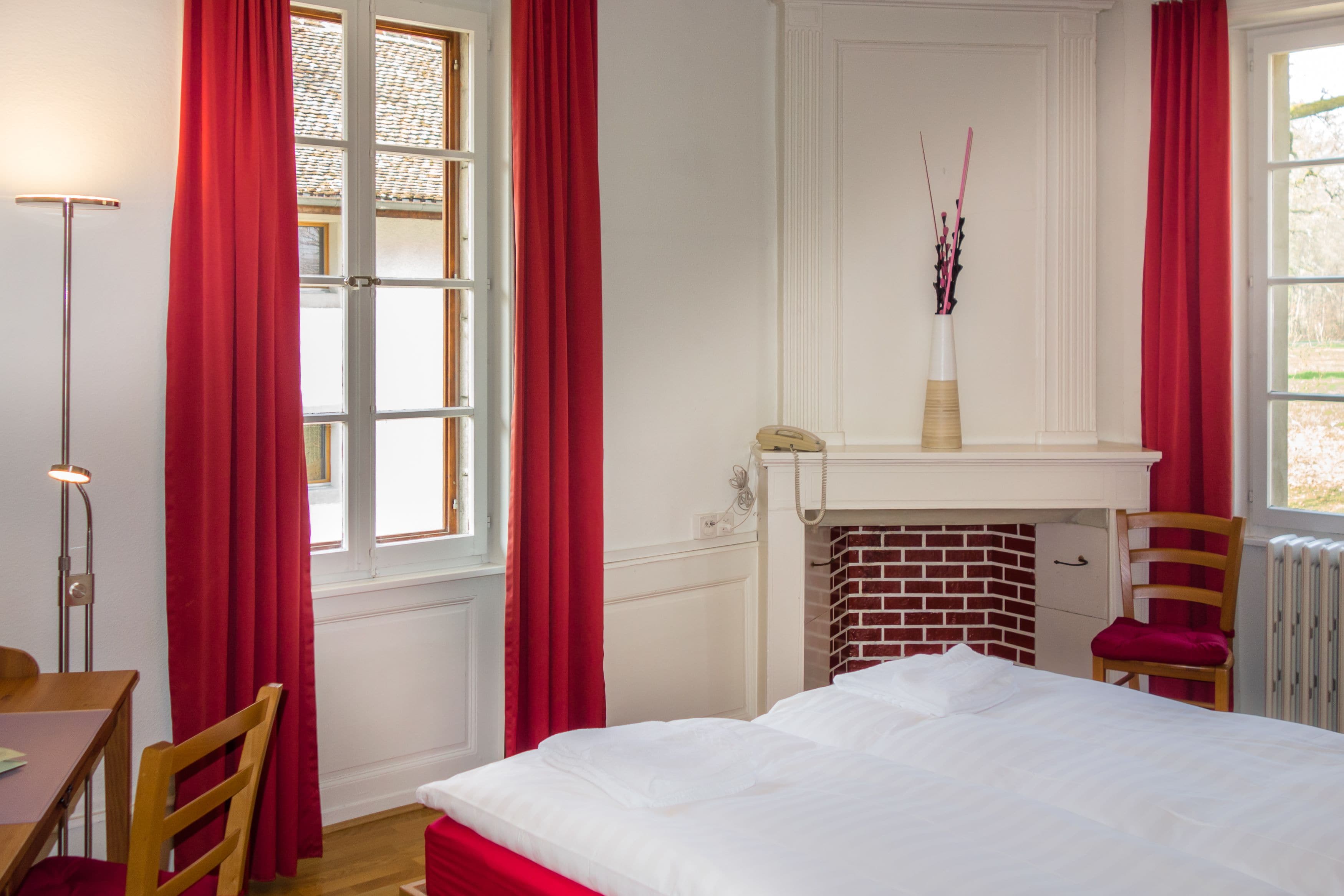 Een slaapkamer met een bed, een stoel, een bureau, een lamp, een open haard en een raam met rode gordijnen.