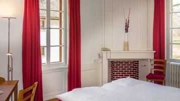 Un dormitorio con una cama, una silla, un escritorio, una lámpara, una chimenea y una ventana con cortinas rojas.