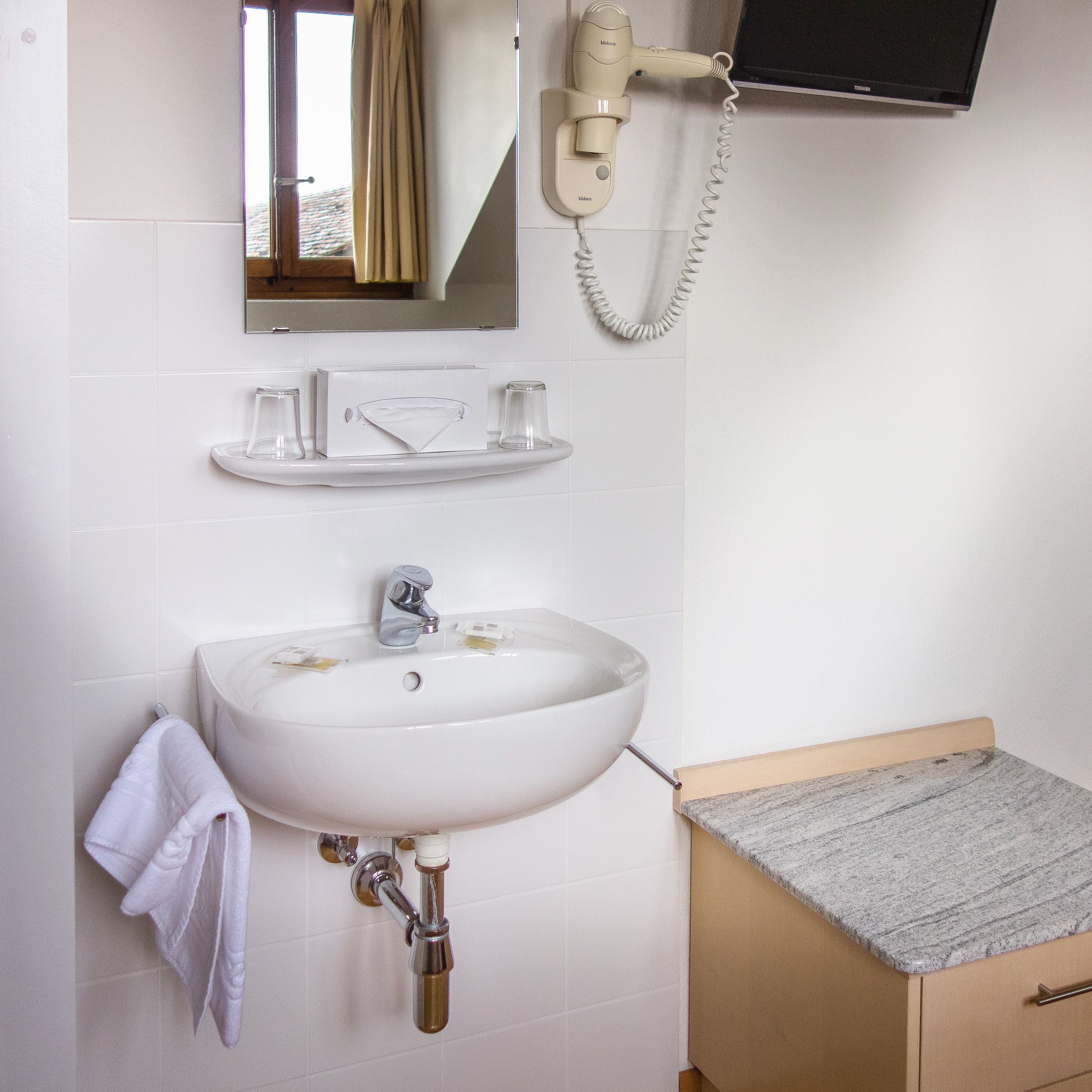 Een badkamer met een wit wastafelmeubel, een haardroger, een spiegel en een televisie die aan de muur is bevestigd.