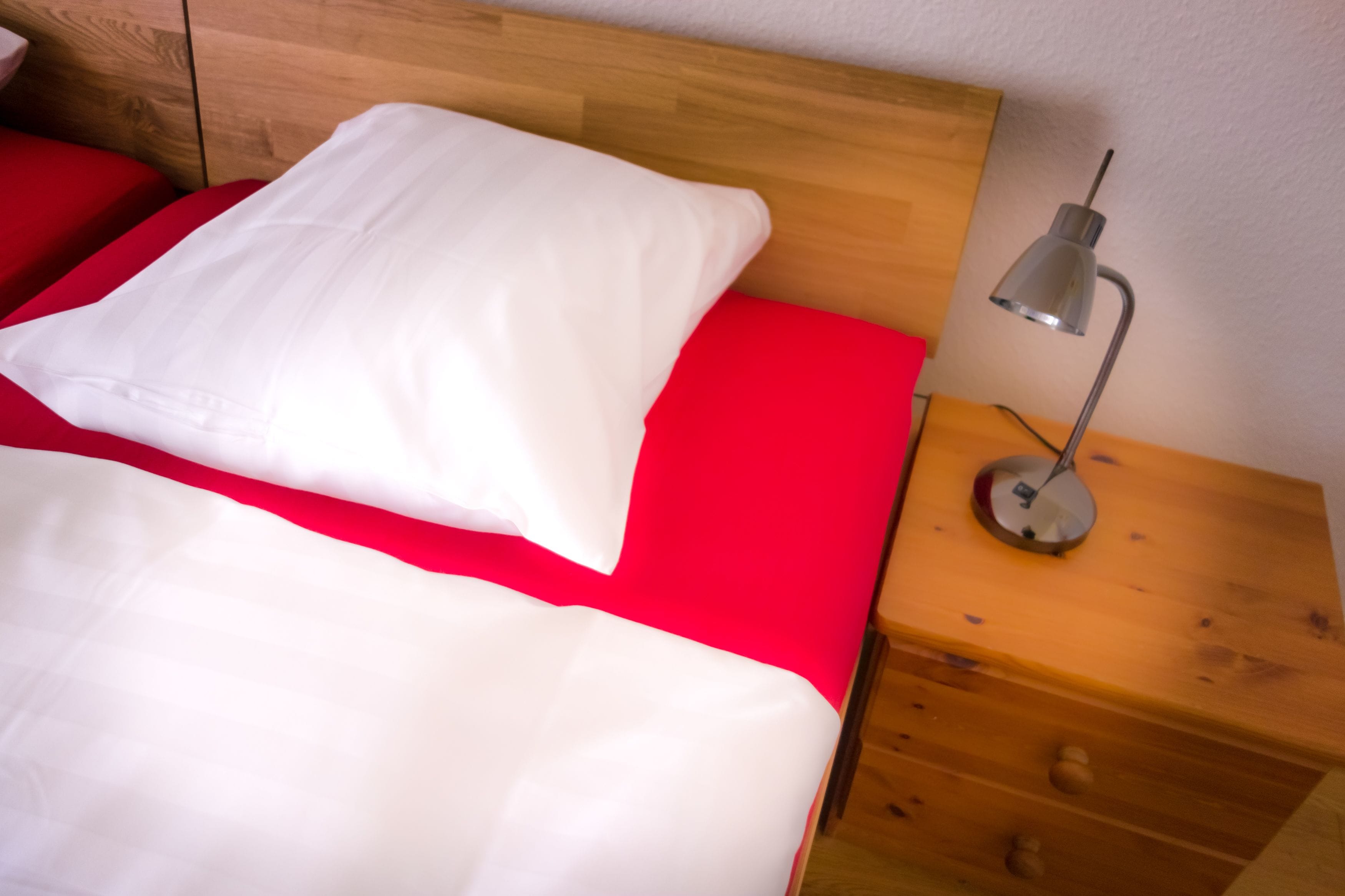 A bed with white and red bedding, a wooden headboard, and a bedside table with a lamp.