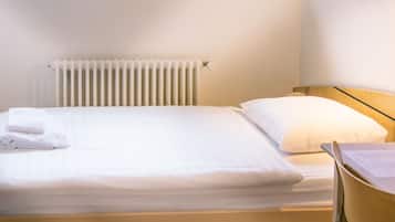 A neatly made bed with white linens, a bedside table with a remote control and papers, and a radiator on the wall.