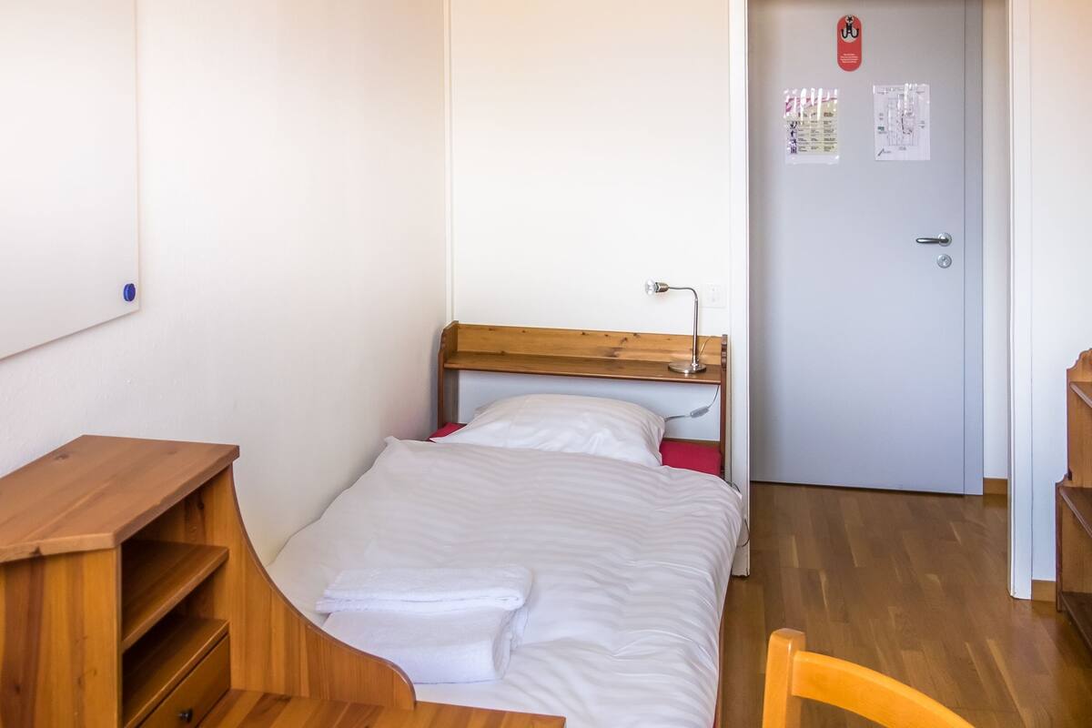 Ein kleiner, ordentlicher Raum mit einem Bett, einem Schreibtisch und einem Stuhl. Neben dem Bett steht eine Holzregal-Konstruktion und eine Tür mit einer Karte und einem Brandschutznachweis.