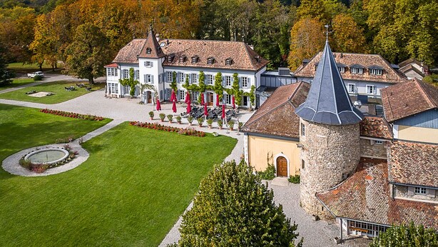 Un grand bâtiment historique, doté d’une tour imposante, entouré de jardins bien entretenus et offrant une vue dégagée sur le paysage environnant.