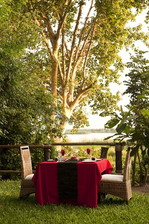 An outdoor dining setup with a table set for two, surrounded by lush greenery and a large tree. - Selous Kinga Lodge (Mloka)