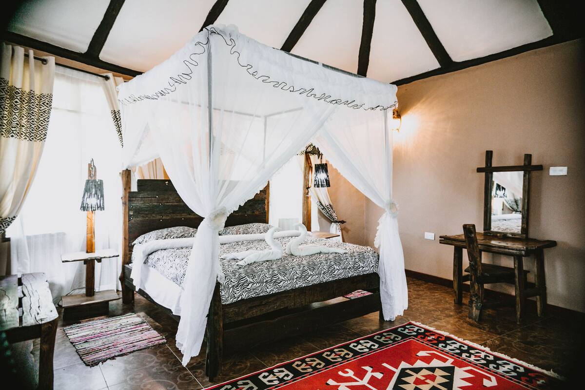 Un dormitorio con cama con dosel, cabecera de madera, alfombra roja estampada y un espejo en la pared.