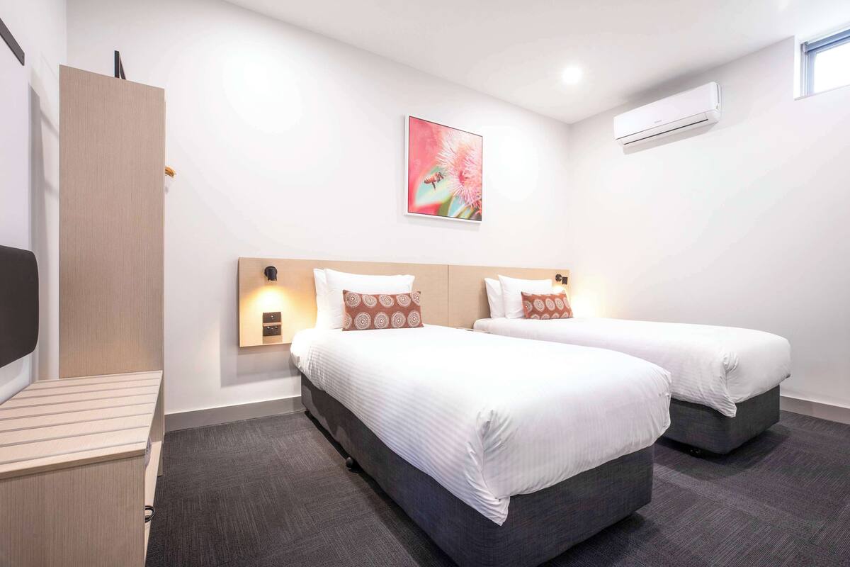 Habitación de hotel con dos camas, un escritorio, aire acondicionado de pared y un cuadro de una flor.
