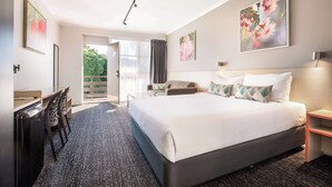 A modern hotel room with a large bed, a desk, and a chair. There is a balcony with a view of greenery. - Nightcap at Ferntree Gully Hotel Motel (Ferntree Gully)