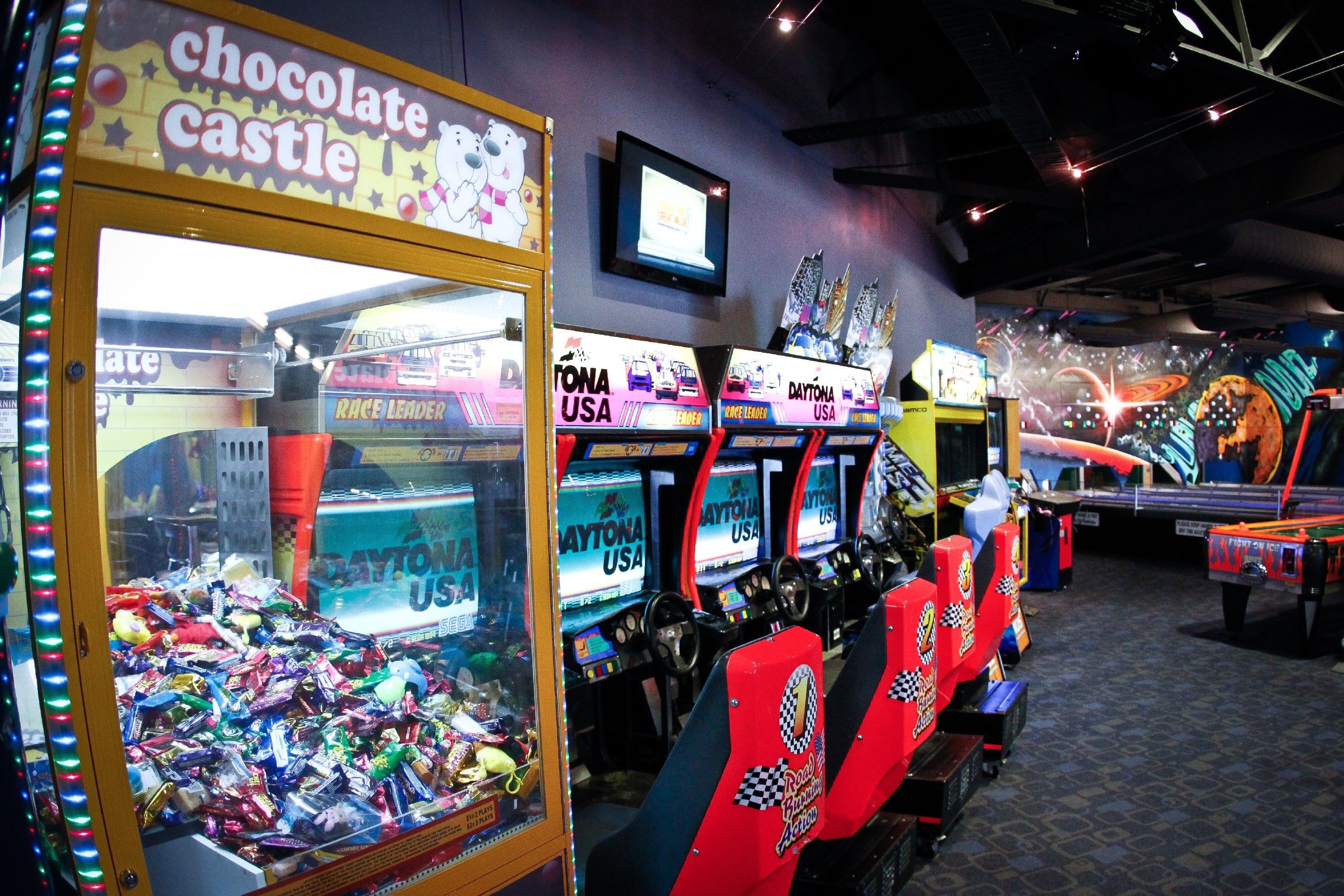A row of racing game machines with "Daytona USA" branding, a "chocolate castle" claw machine, and a television screen on the wall.