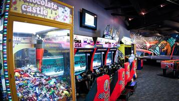 A row of racing game machines with "Daytona USA" branding, a "chocolate castle" claw machine, and a television screen on the wall.