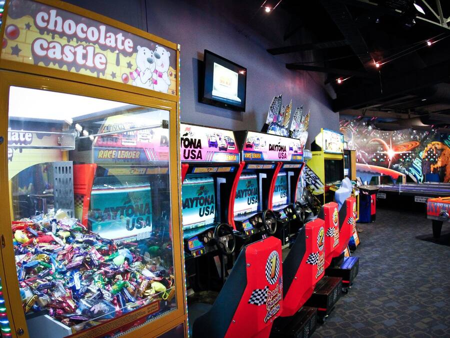 A row of racing game machines with "Daytona USA" branding, a "chocolate castle" claw machine, and a television screen on the wall.