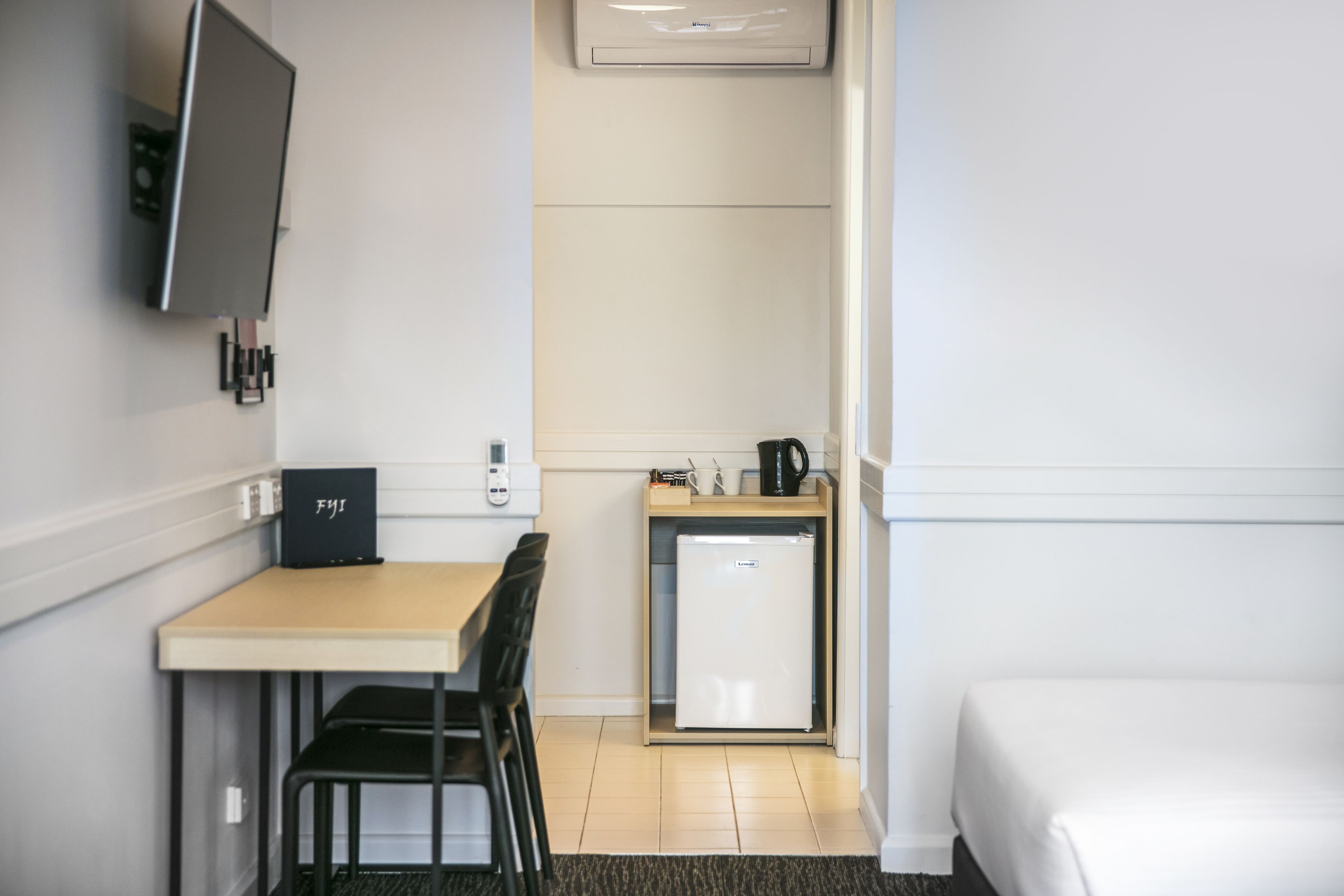 Een compacte hotelkamer met een bed, een kleine kitchenette met een koelkast en koffiezetapparaat, een bureau met stoel, en een aan de muur bevestigde televisie.