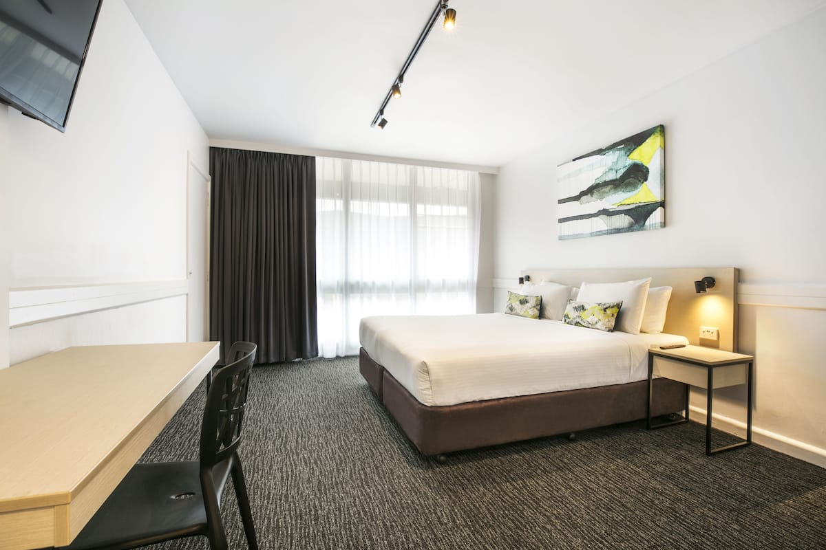 Quarto de hotel com cama, escrivaninha, cadeira, mesa de cabeceira e um quadro na parede.