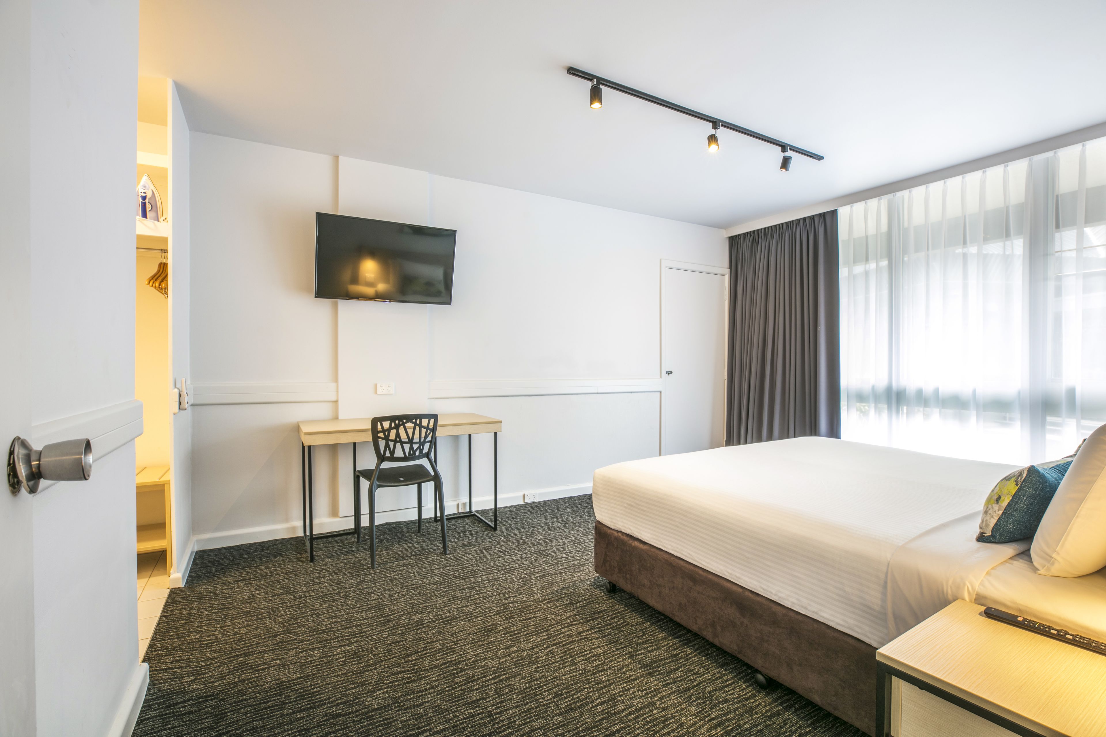 A hotel room with a bed, a desk, a chair, a TV mounted on the wall, and a large window with curtains.