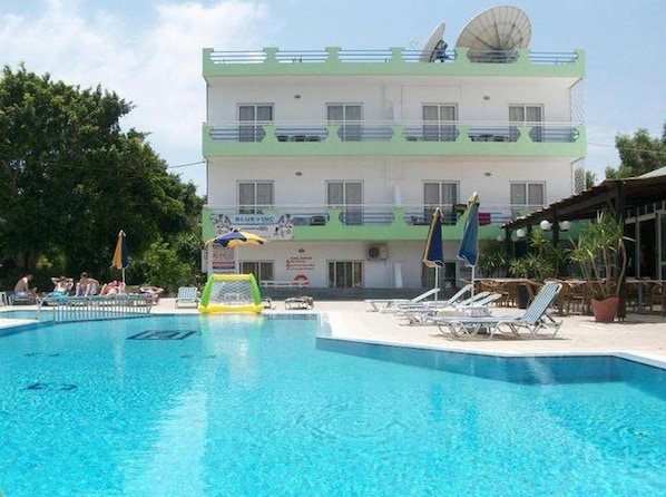 A swimming pool with lounge chairs and umbrellas, a building with multiple balconies, and a sign for 'BLUE INC'.