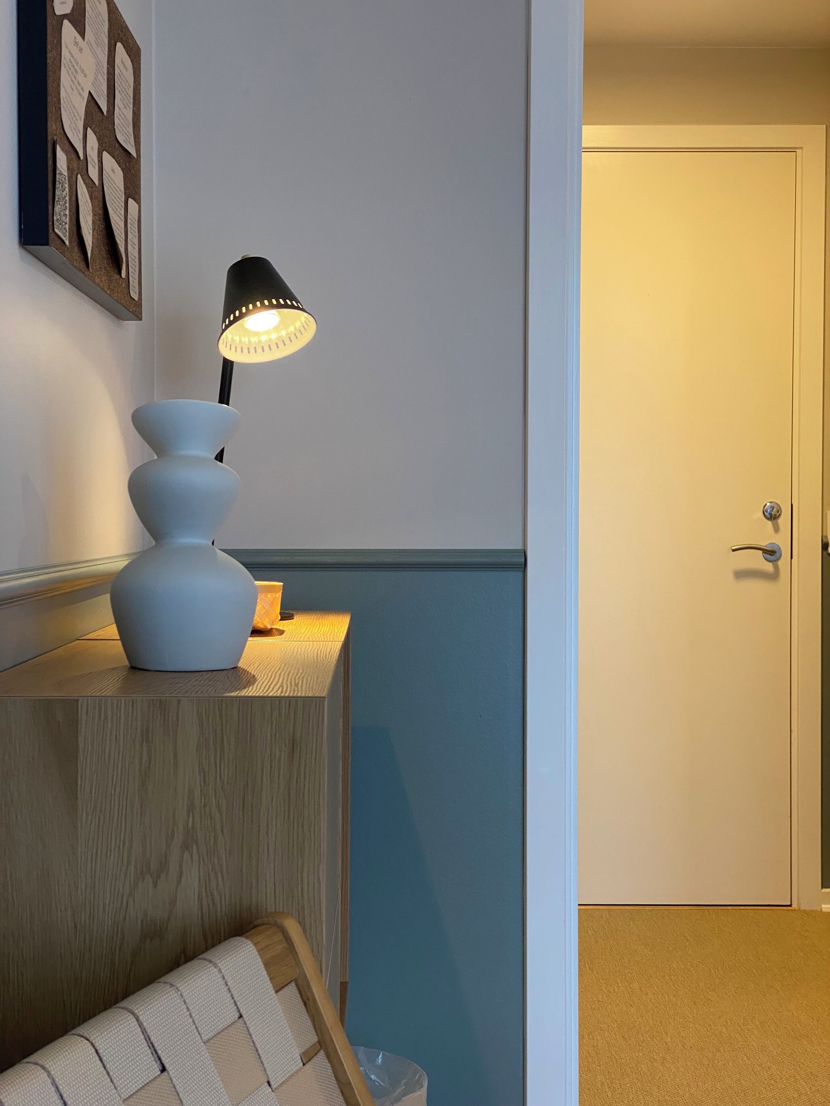 A room with a wooden console table, a white vase, and a black lamp. There is a door to the right and a wall with a framed picture.
