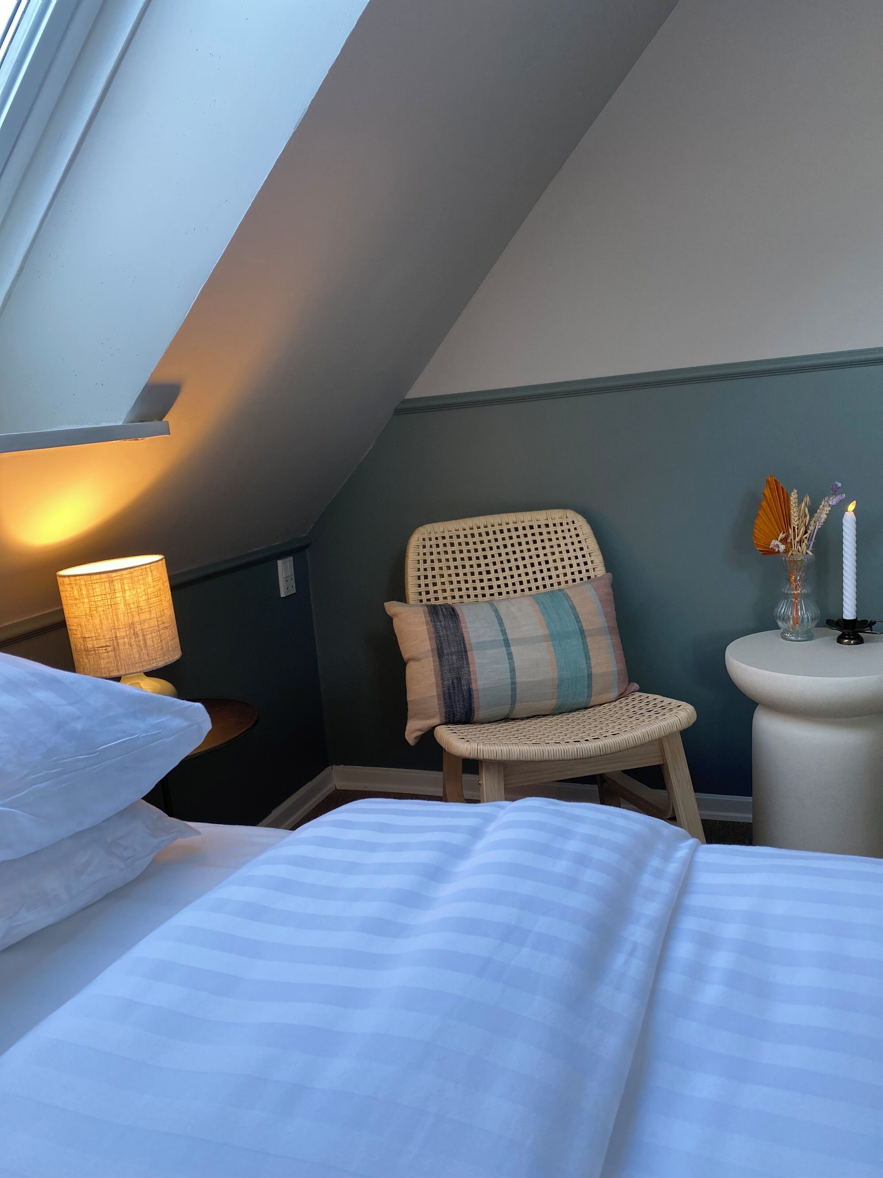 A cozy bedroom with a sloped ceiling, a wicker chair with a striped pillow, a bedside table with a lamp, and a small vase with candles.