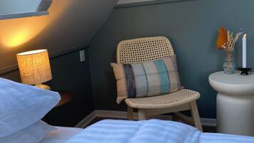 A cozy bedroom with a sloped ceiling, a wicker chair with a striped pillow, a bedside table with a lamp, and a small vase with candles.