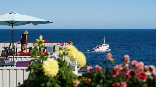 A coastal scene with a boat, a woman, and a patio with flowers and umbrellas. - Hotel Klippen  (Gudhjem)