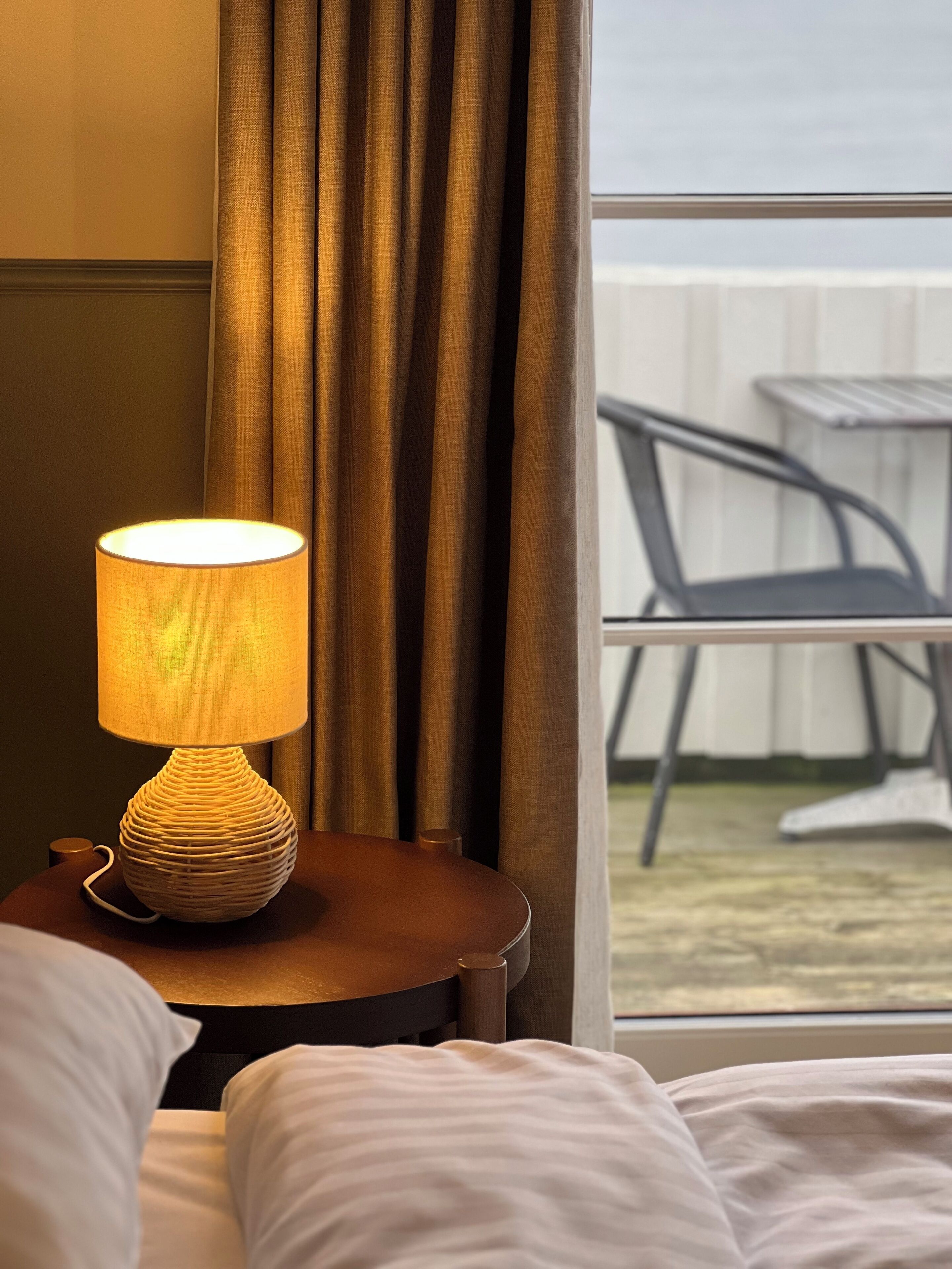 Une chambre d’hôtel avec un lit, une table de chevet avec une lampe, une fenêtre avec des rideaux et une vue sur des meubles d’extérieur.