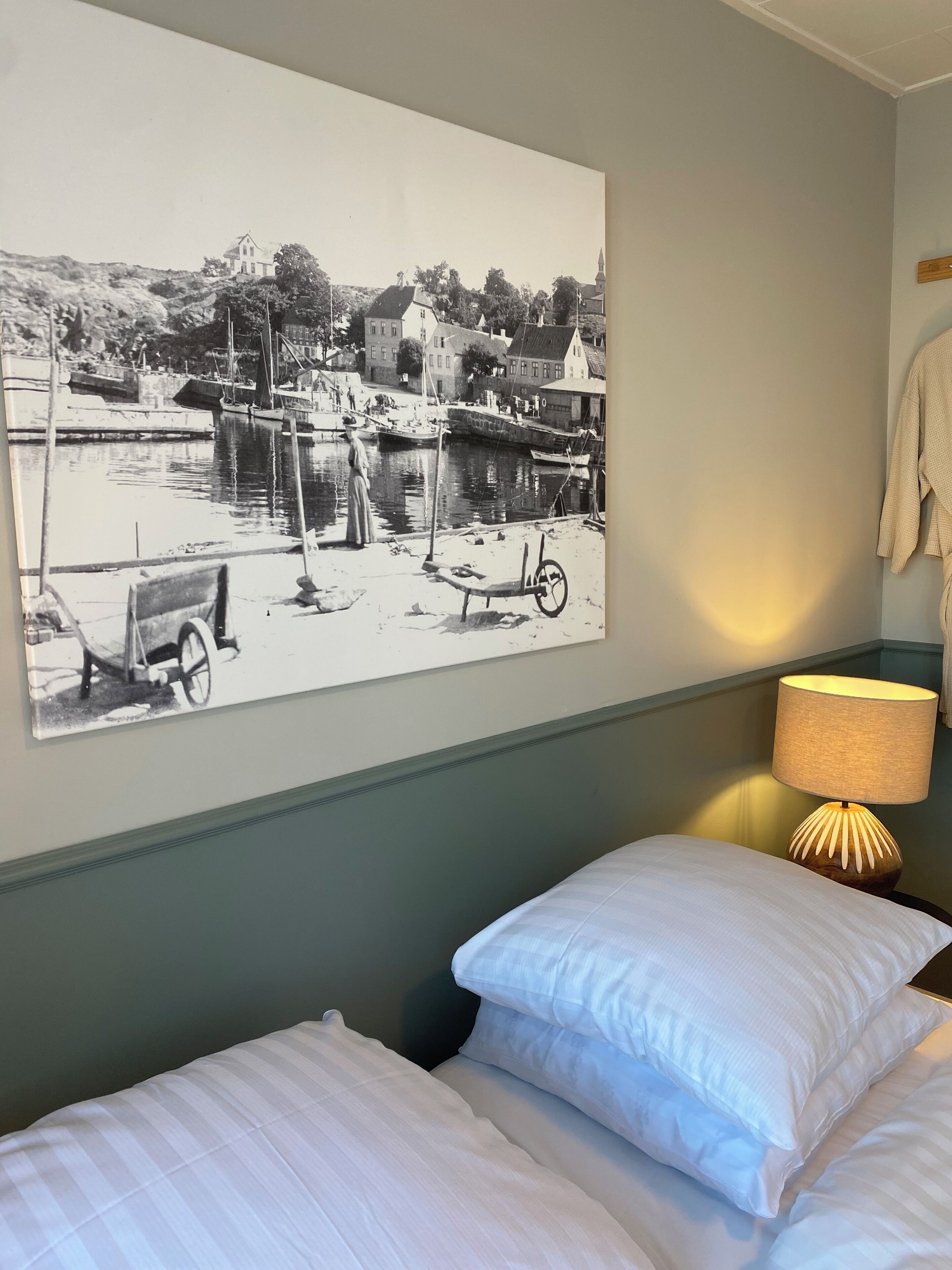 Un dormitorio con cama, almohadas, una lámpara de pared y una fotografía grande en blanco y negro de una escena junto al agua.