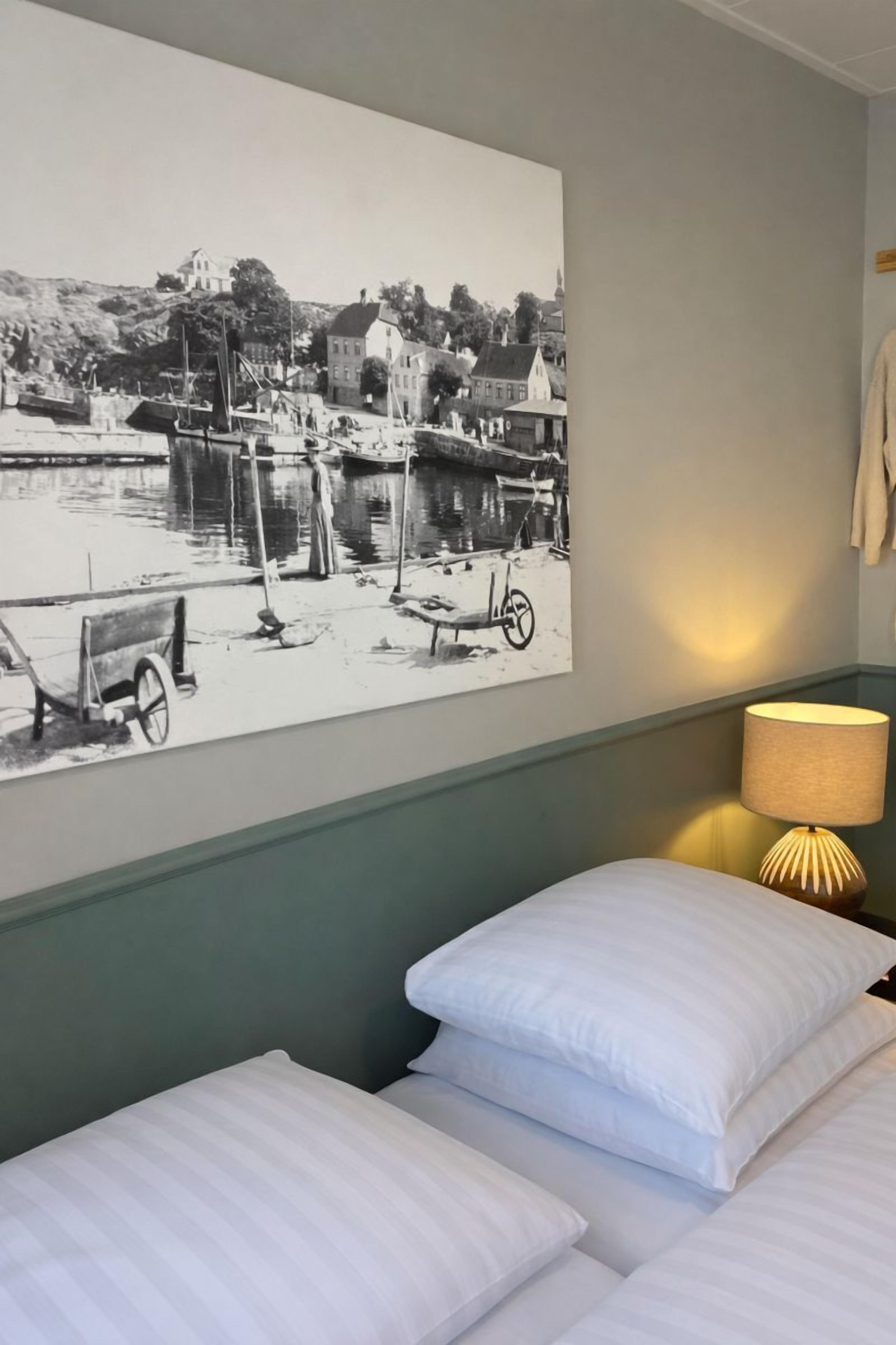 A hotel room with a bed, pillows, a wall-mounted lamp, and a black and white photograph of a waterfront scene.