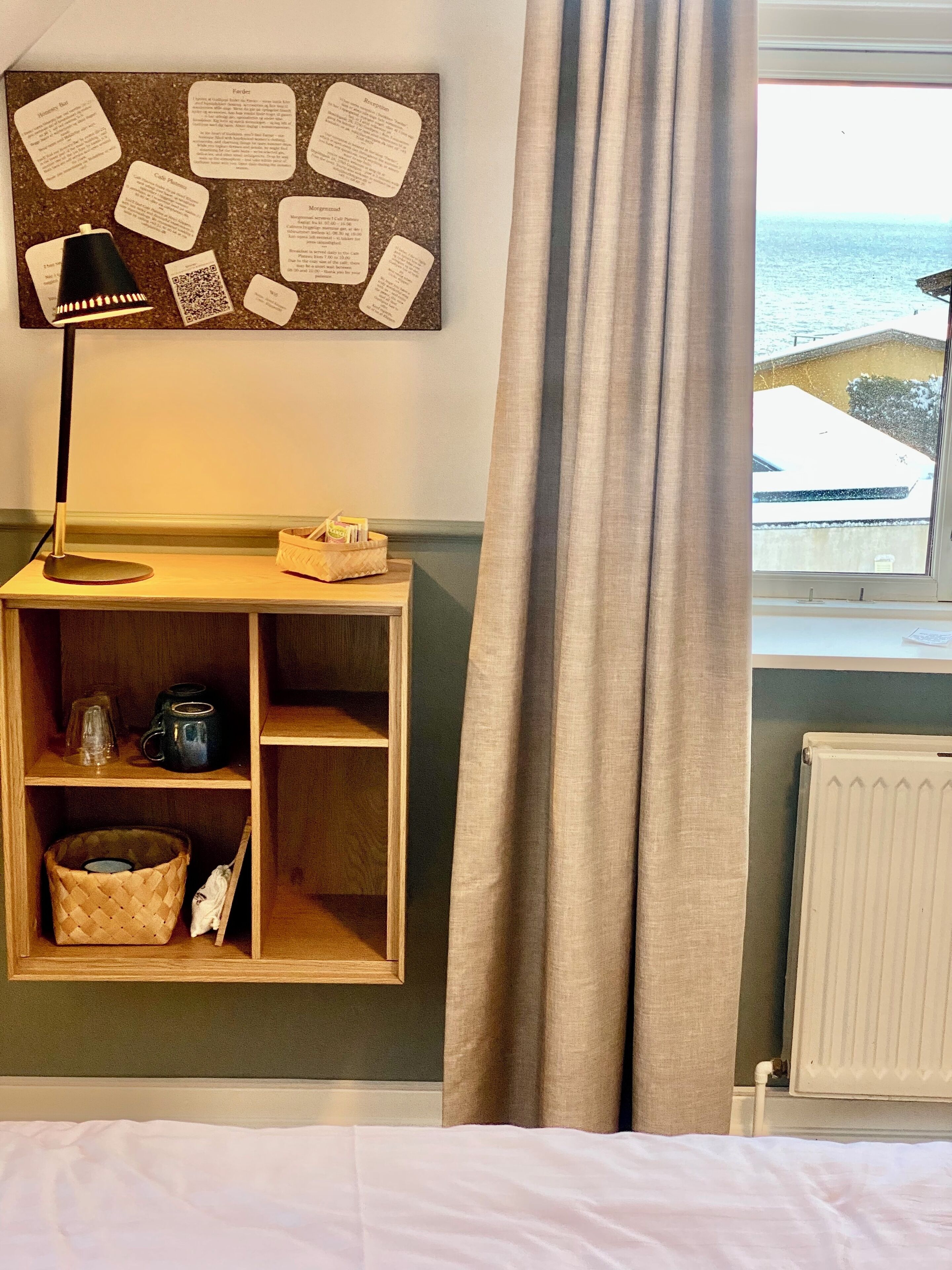 A room with a bulletin board, a lamp, a shelf with mugs, a basket, and a view of the sea through a window.