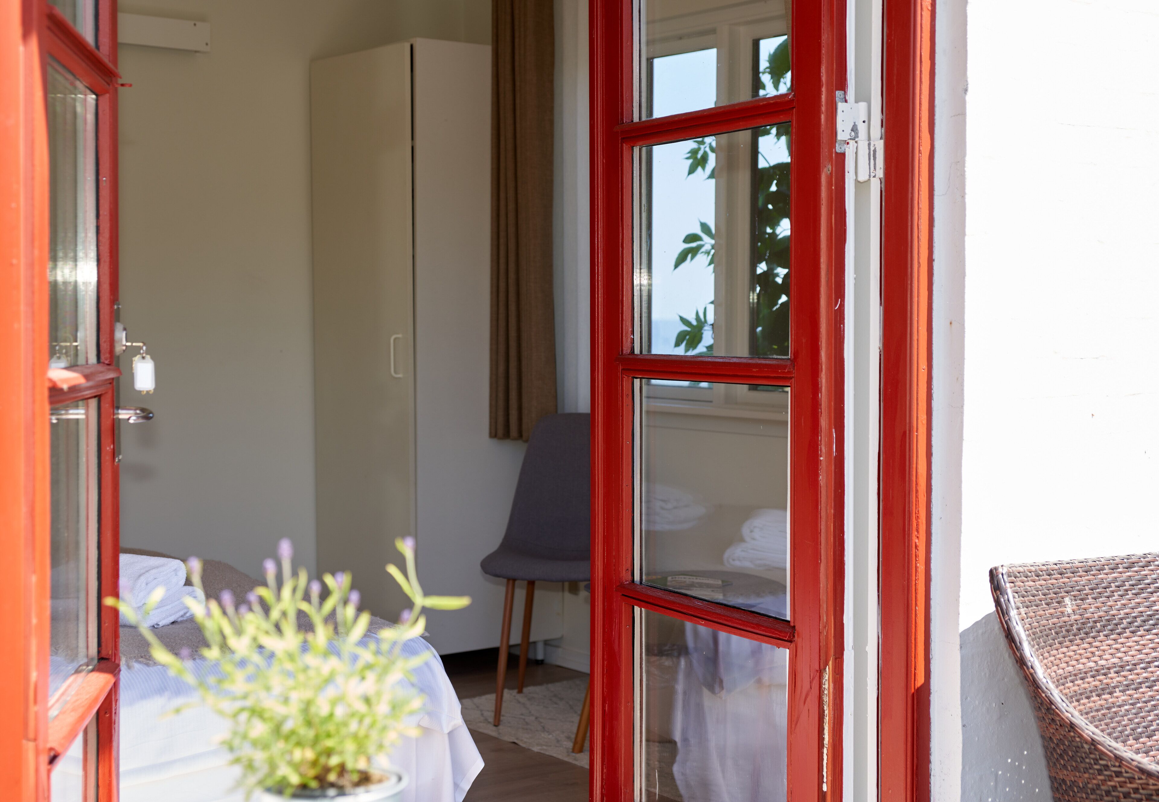 Quarto com cama, cadeira, mesa com planta e janela com vista para árvores.