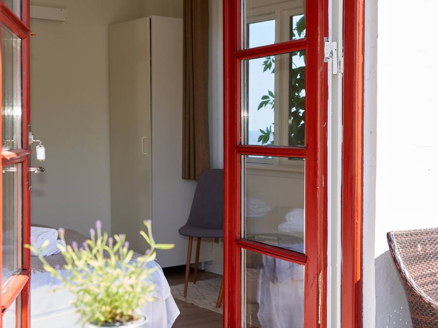 Quarto com cama, cadeira, mesa com planta e janela com vista para árvores.