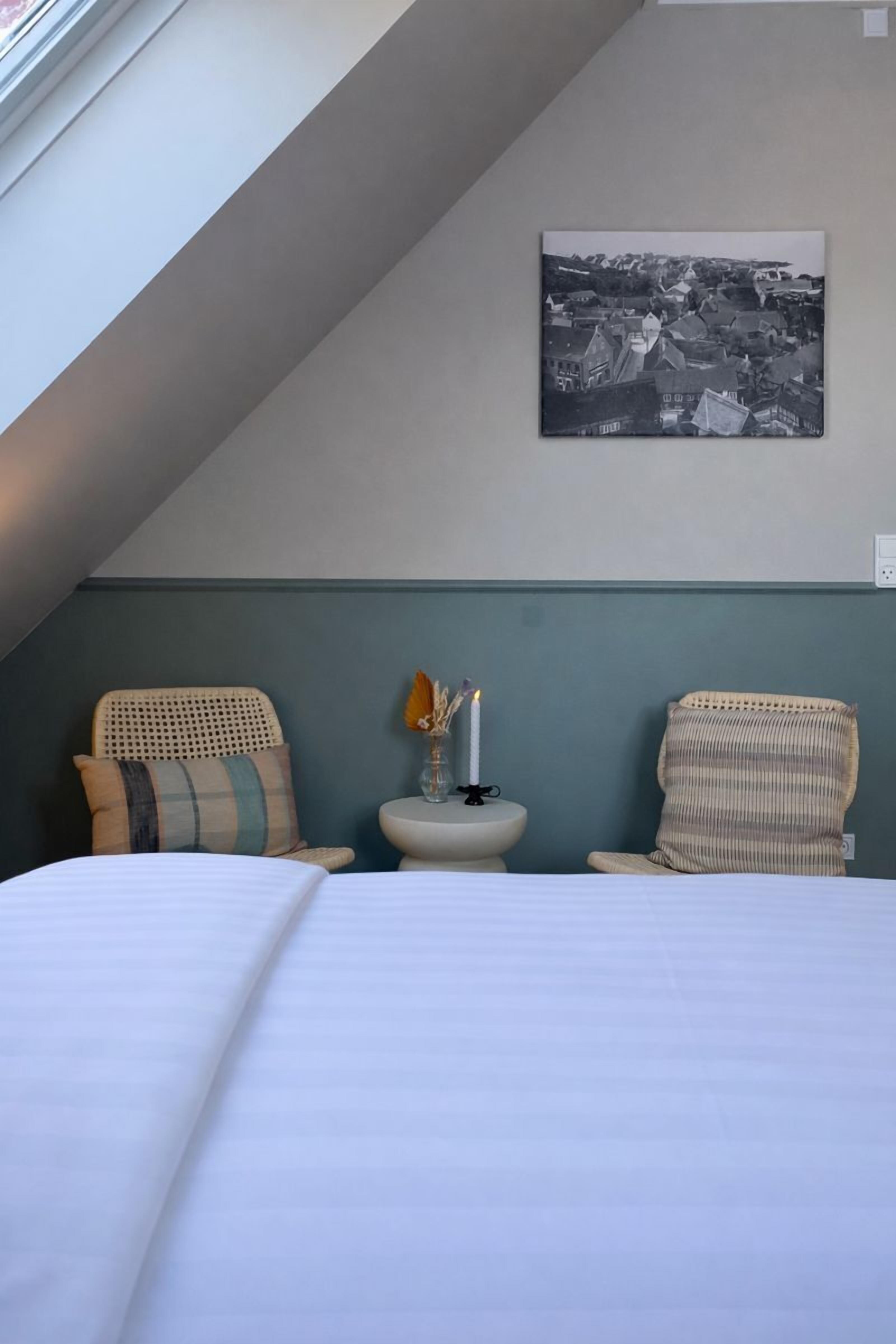 A bedroom with a large bed, two wicker chairs, a small bedside table with a candle, and a black and white cityscape painting on the wall.