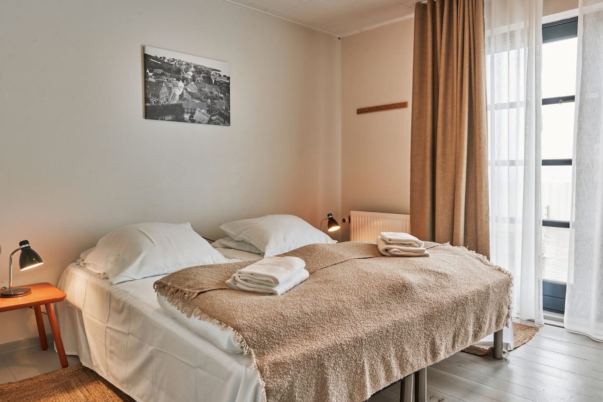 A neatly made bed with a blanket, pillows, and folded towels. There is a small bedside table with a lamp, a radiator, and a window with curtains.