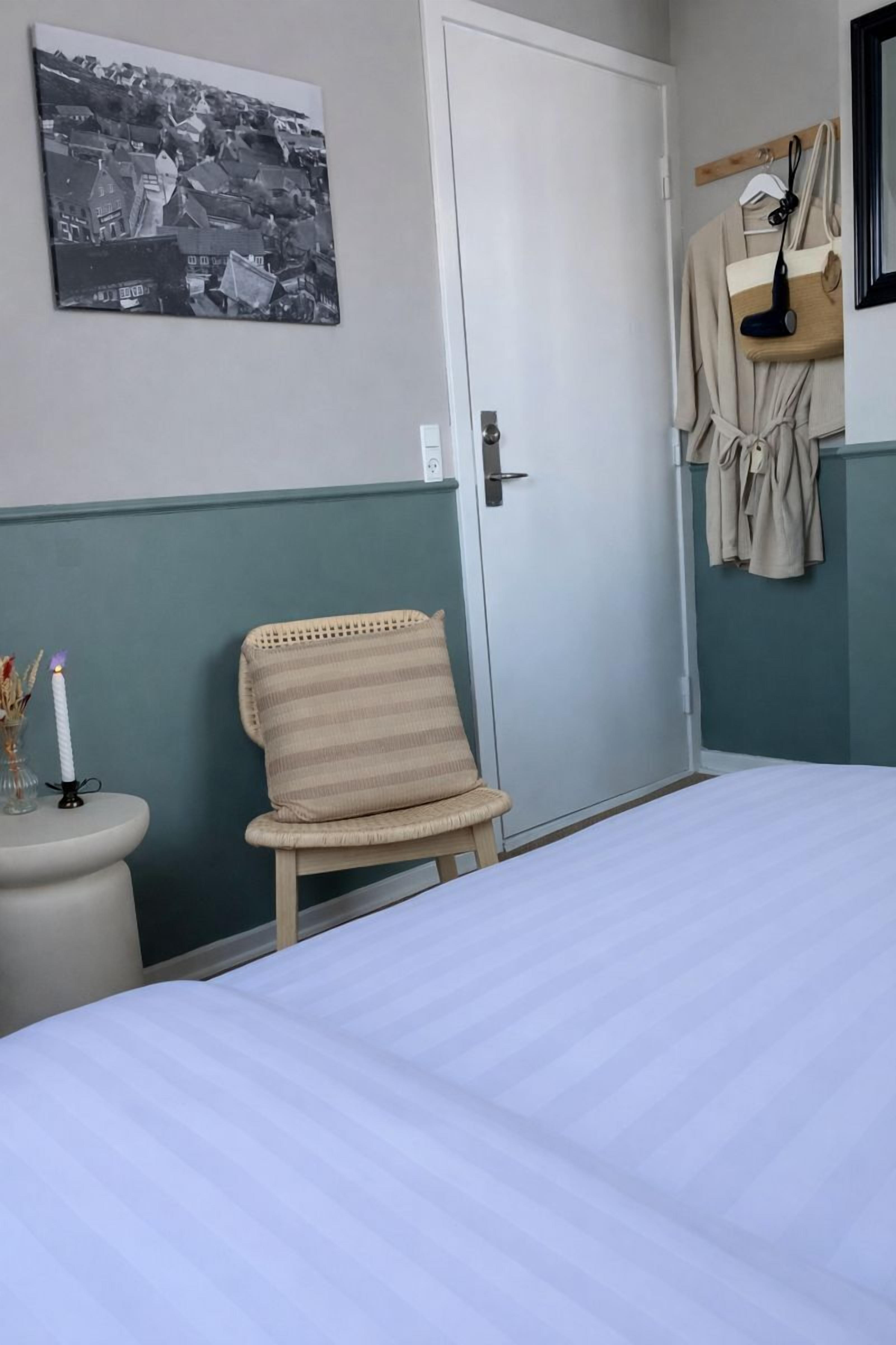 A bedroom with a bed, a chair, a bedside table with a candle, a coat rack, and a framed picture on the wall.