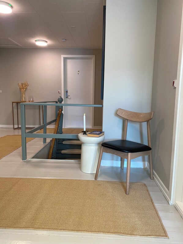 Un pasillo con barandilla azul, un pedestal blanco, una silla de madera y una puerta con el número 7.