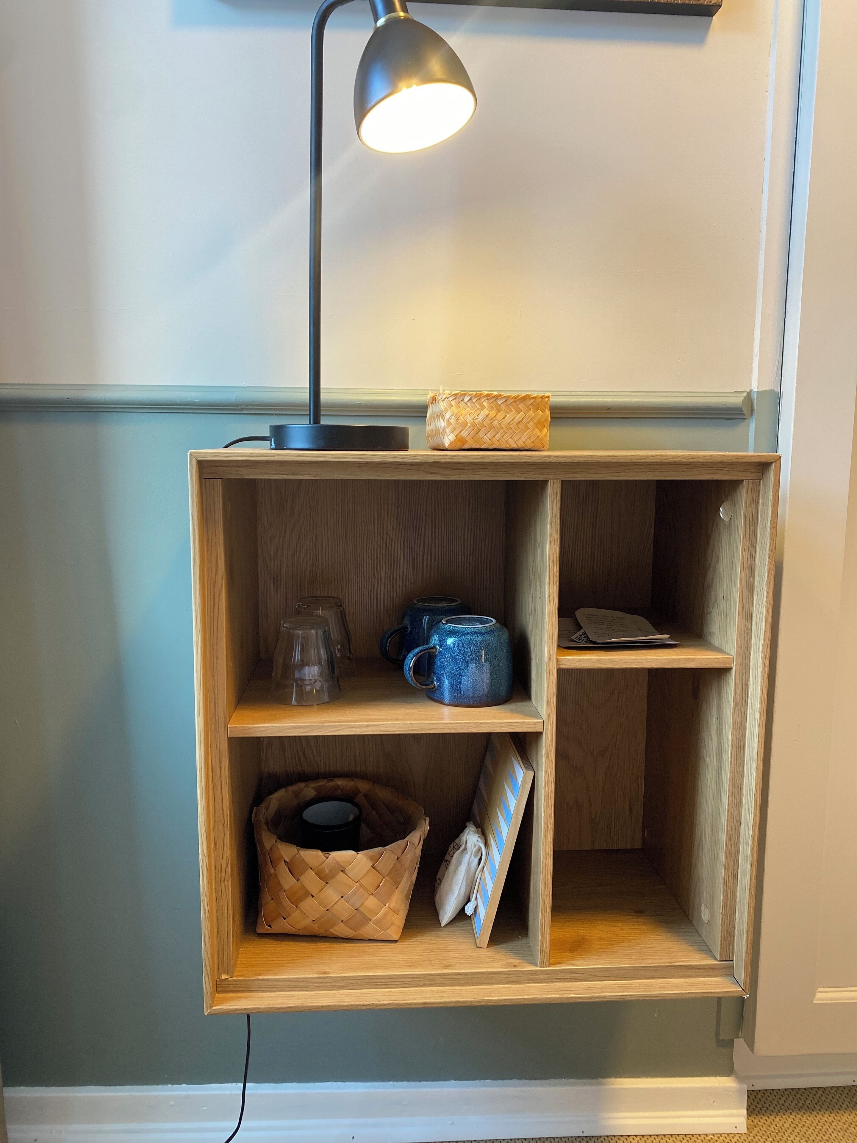 Una estantería de madera con una lámpara de escritorio negra, una cesta tejida y una taza azul.