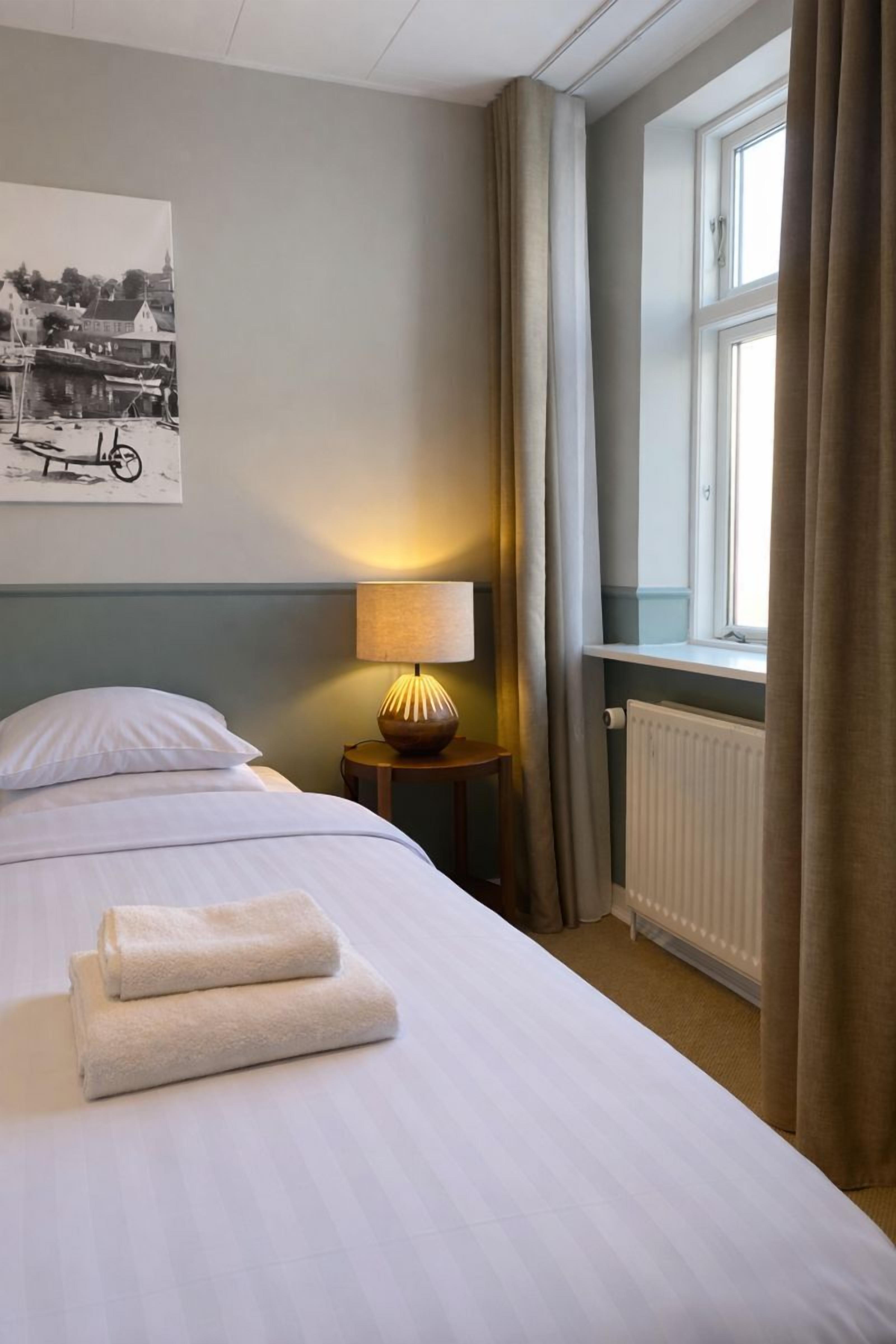 A hotel room with a neatly made bed, a lamp on a bedside table, a window with curtains, and a poster on the wall.