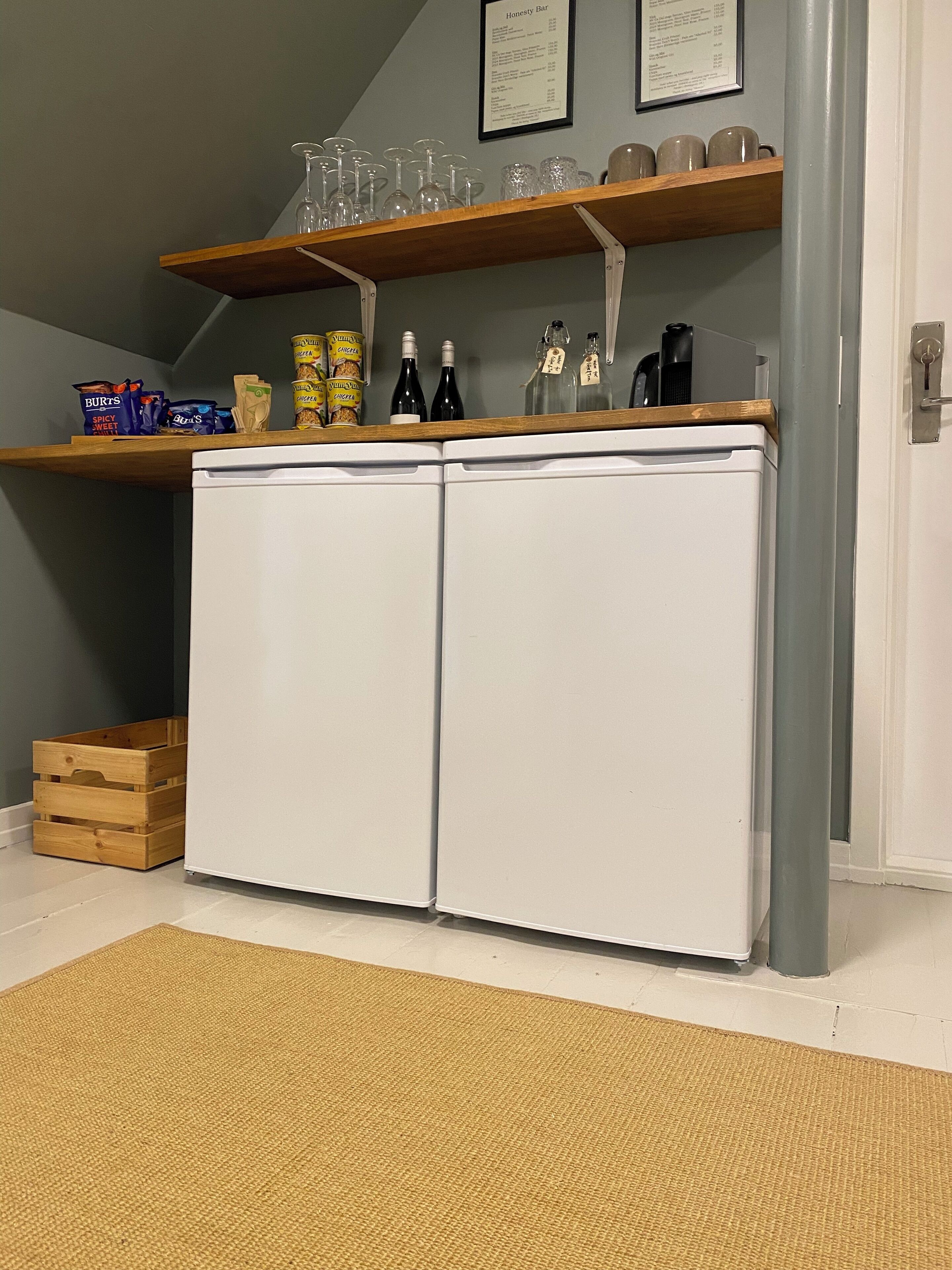 A compact kitchen with a white refrigerator, wooden shelves above holding glassware and condiments, and a small wooden crate.