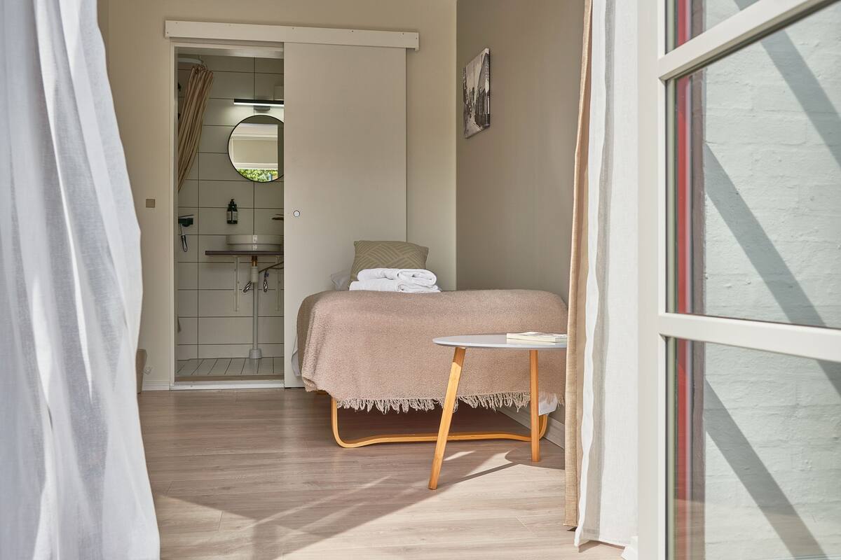 A bedroom with a bed, a small table, a bathroom with a shower, and a window.