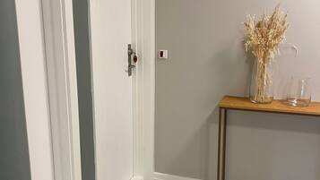 Un pasillo con una puerta cerrada, una consola de madera con un jarrón de flores secas y un interruptor de luz en la pared.