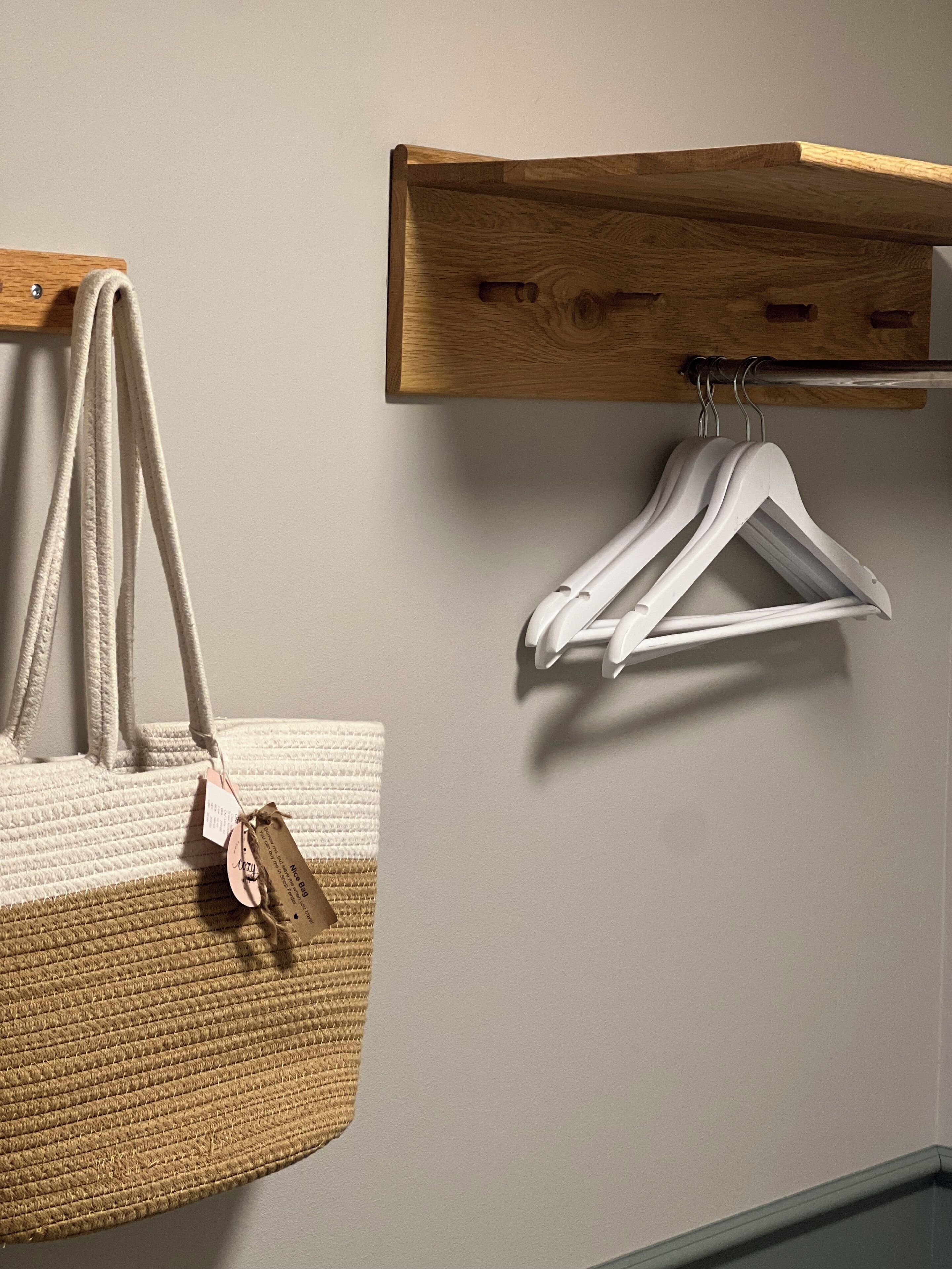 Una habitación con un estante de madera sobre una pared blanca, donde se ven perchas blancas y un bolso tejido con una etiqueta.