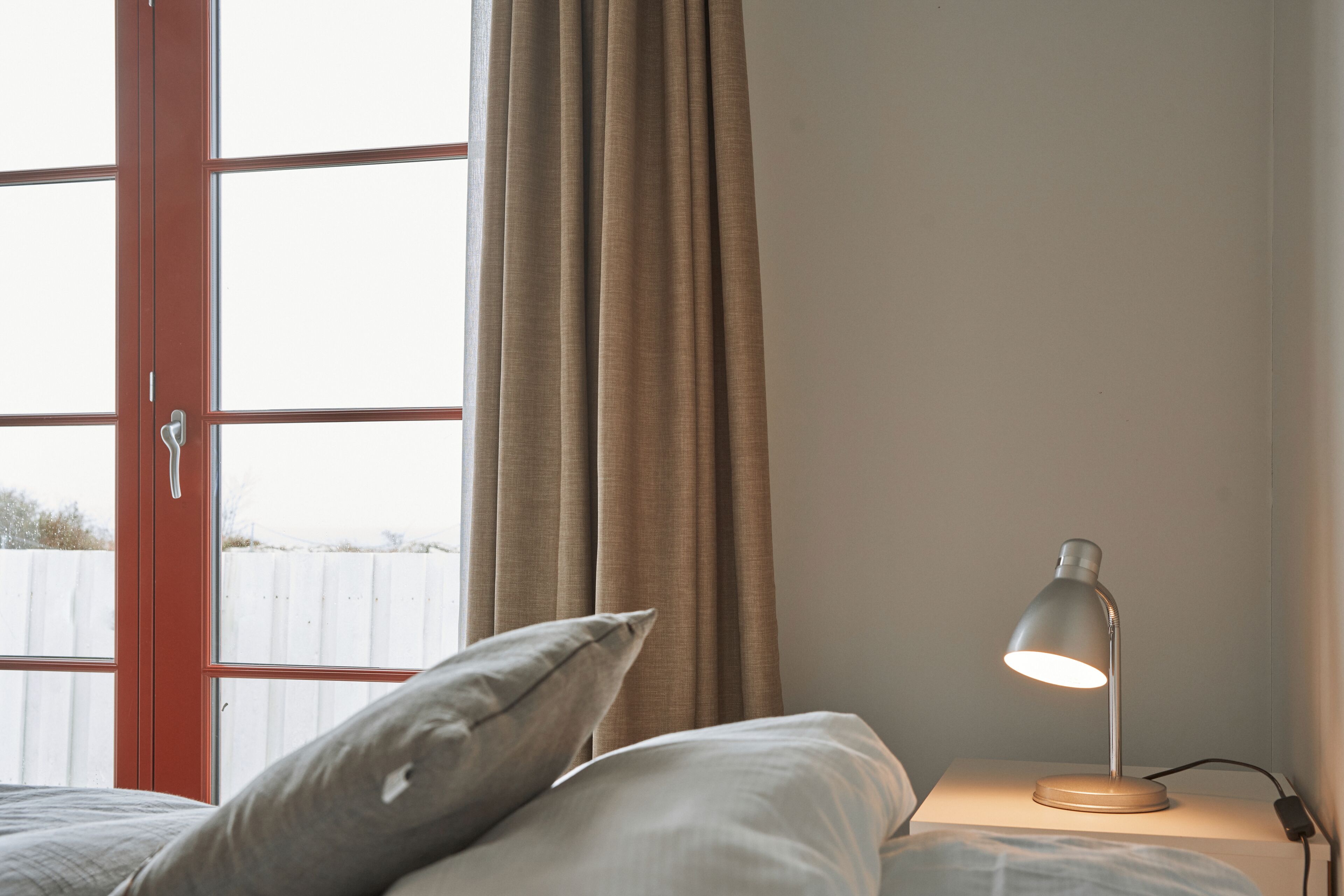 A bedroom with a bed, a bedside table with a lamp, a window with curtains, and a view of a fence outside.